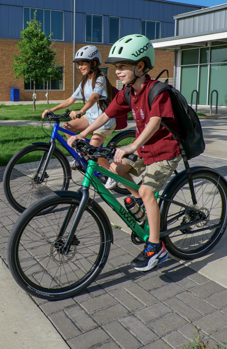 A group of young bike riders steer their woom EXPLORE bikes through a school parking lot. They are wearing woom READY helmets and riding magnetic blue, neon coral, and jungle green woom EXPLORE bikes.