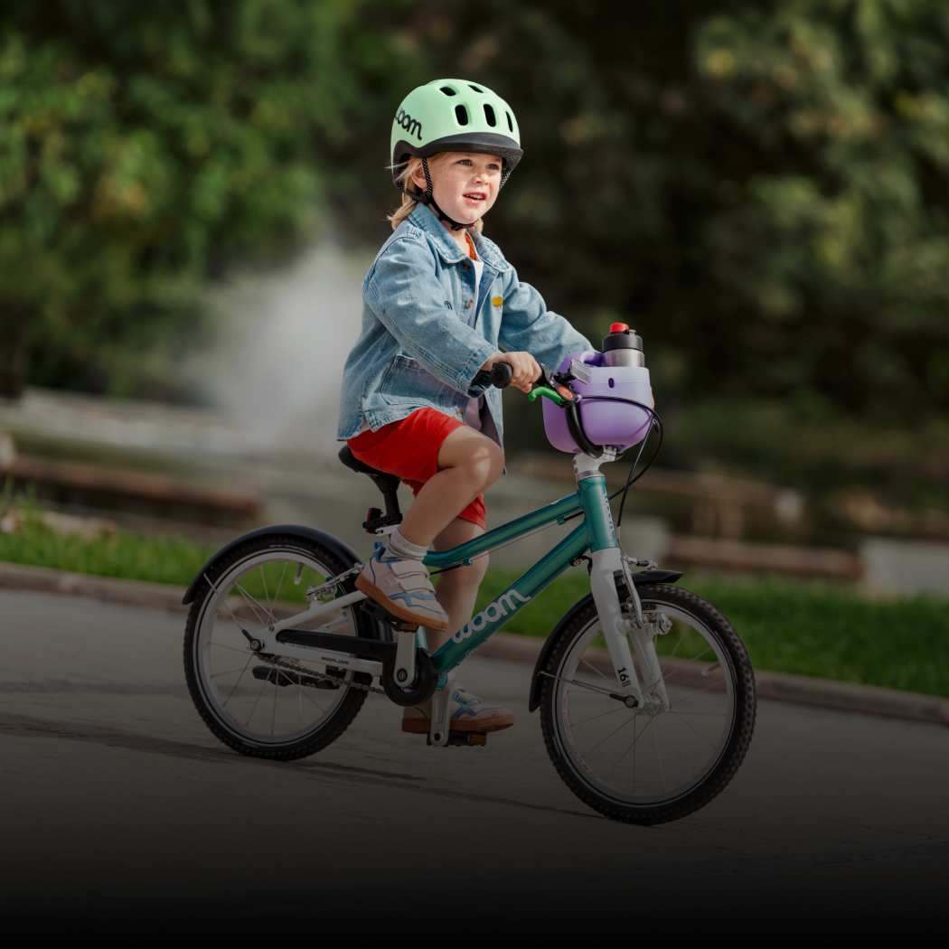 A blonde boy wears a mint READY Helmet while riding his woom GO bike with a POP Basket holding a GLUG Water Bottle attached.