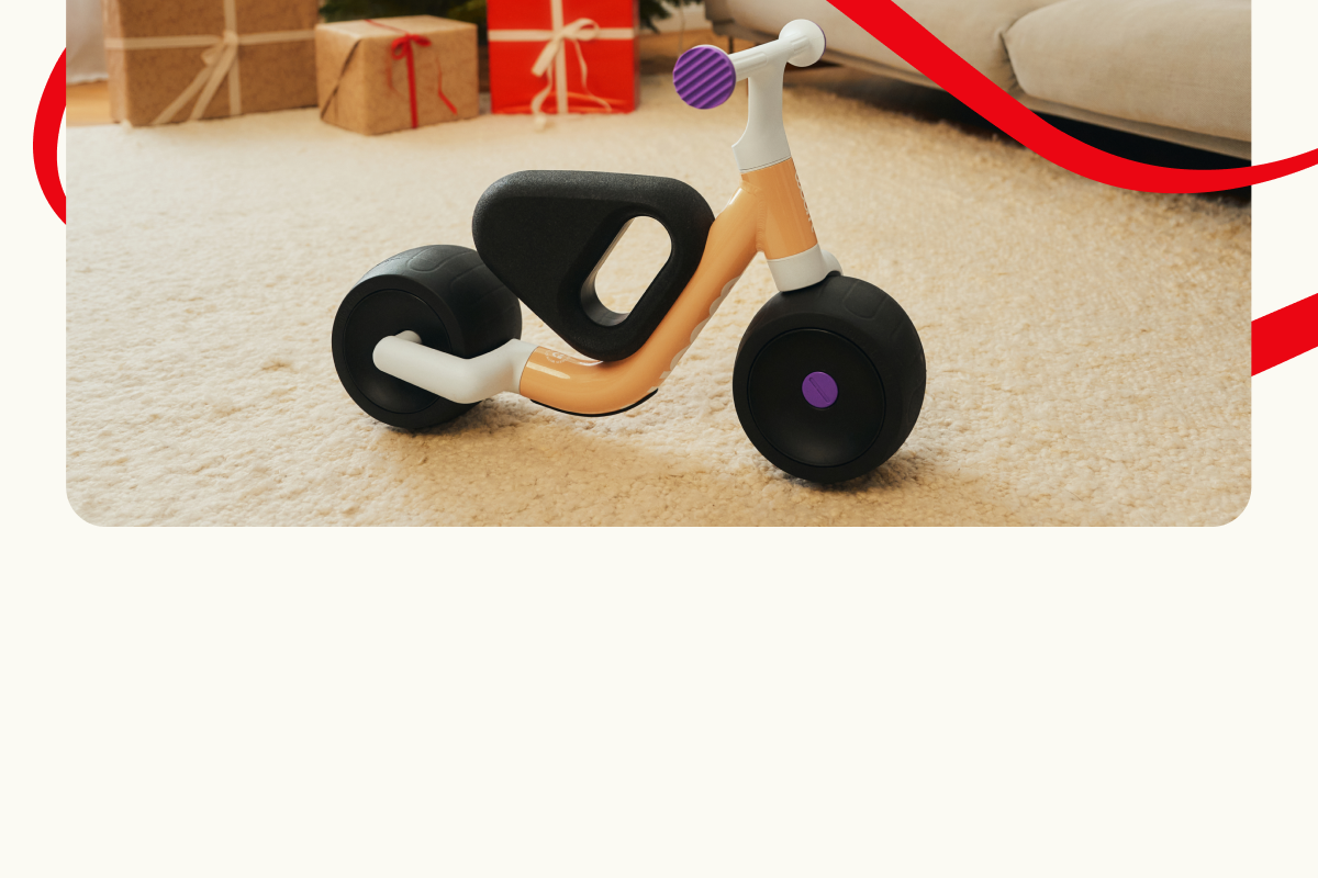 woom WOW drinnen auf einem Teppich, mit Geschenkboxen und einer Couch im Hintergrund. Dekorative rote Linien rahmen die Szene ein.