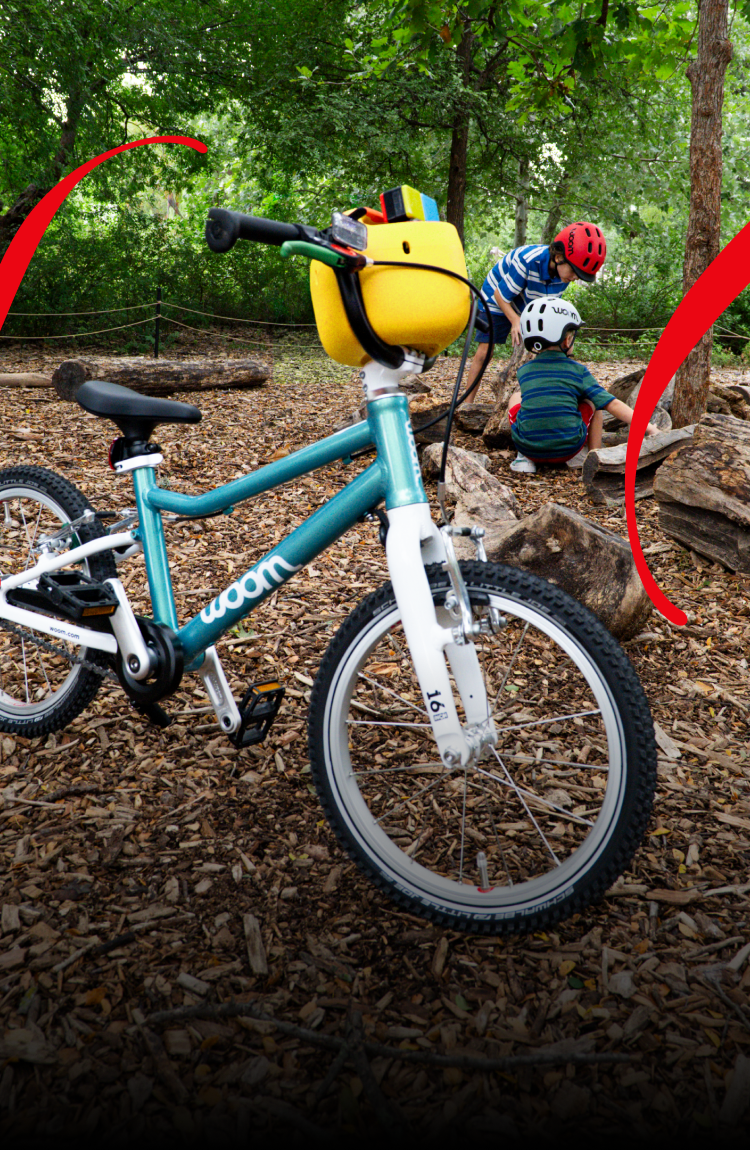 A woom GO in metallic turquoise with a yellow POP bicycle basket on the handlebars stands on a forest floor covered with wood chips, while two children wearing woom READY helmets play in the background.