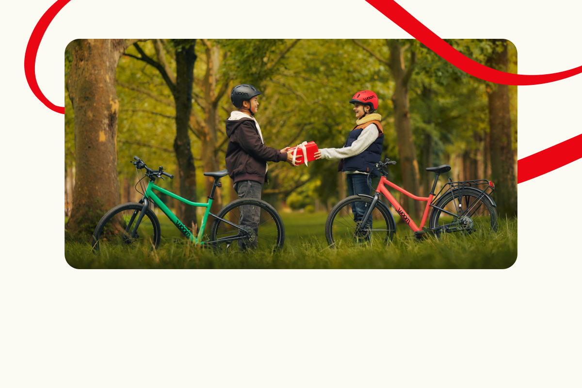 Two kids with woom EXPLORE bikes in a forest are exchanging a gift. Decorative red lines frame the scene.