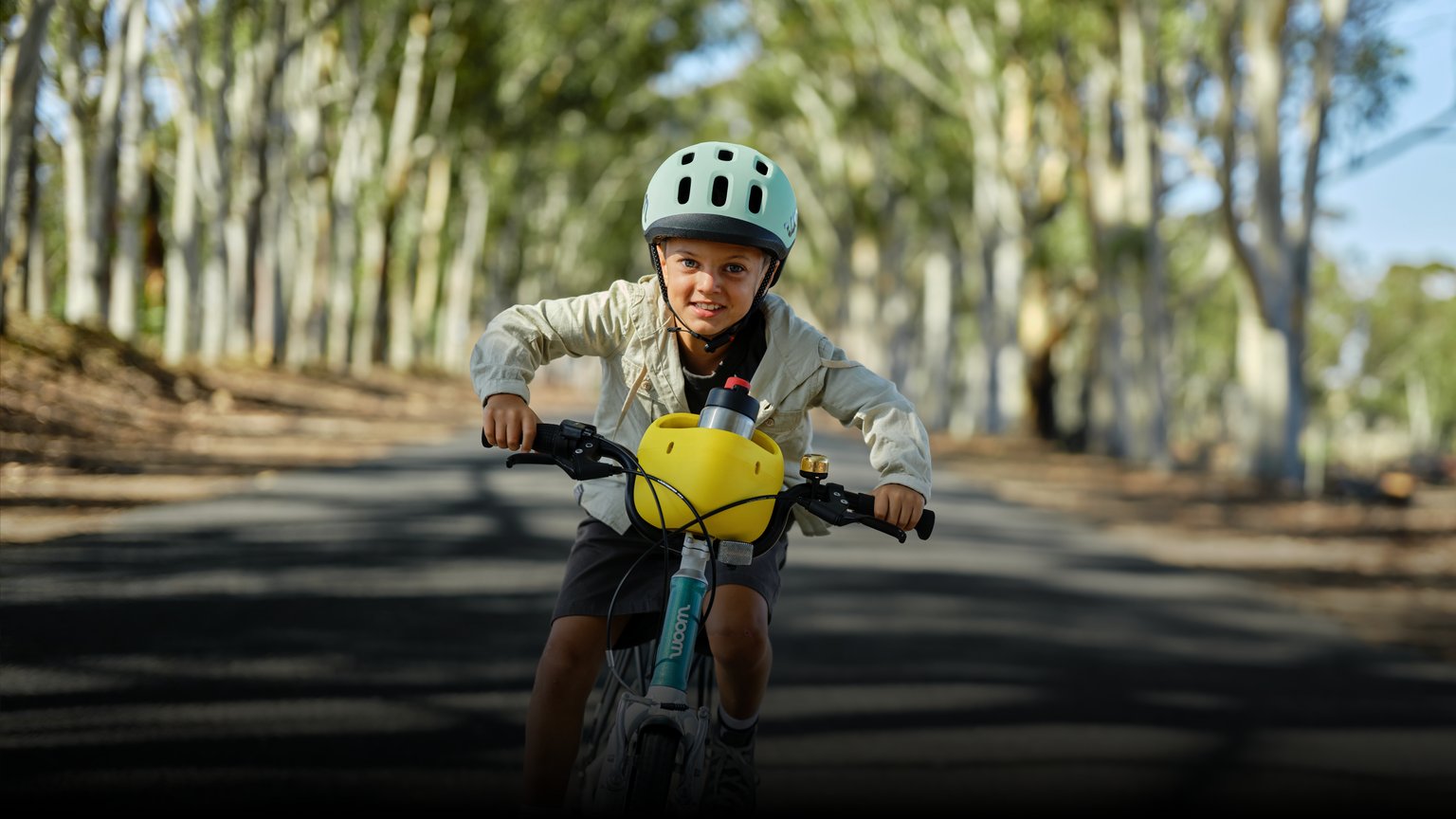 Kind mit READY Helm in Türkis auf dem Kopf fährt mit einem woom GO samt POP Fahrradkorb auf einer Straße im Grünen.