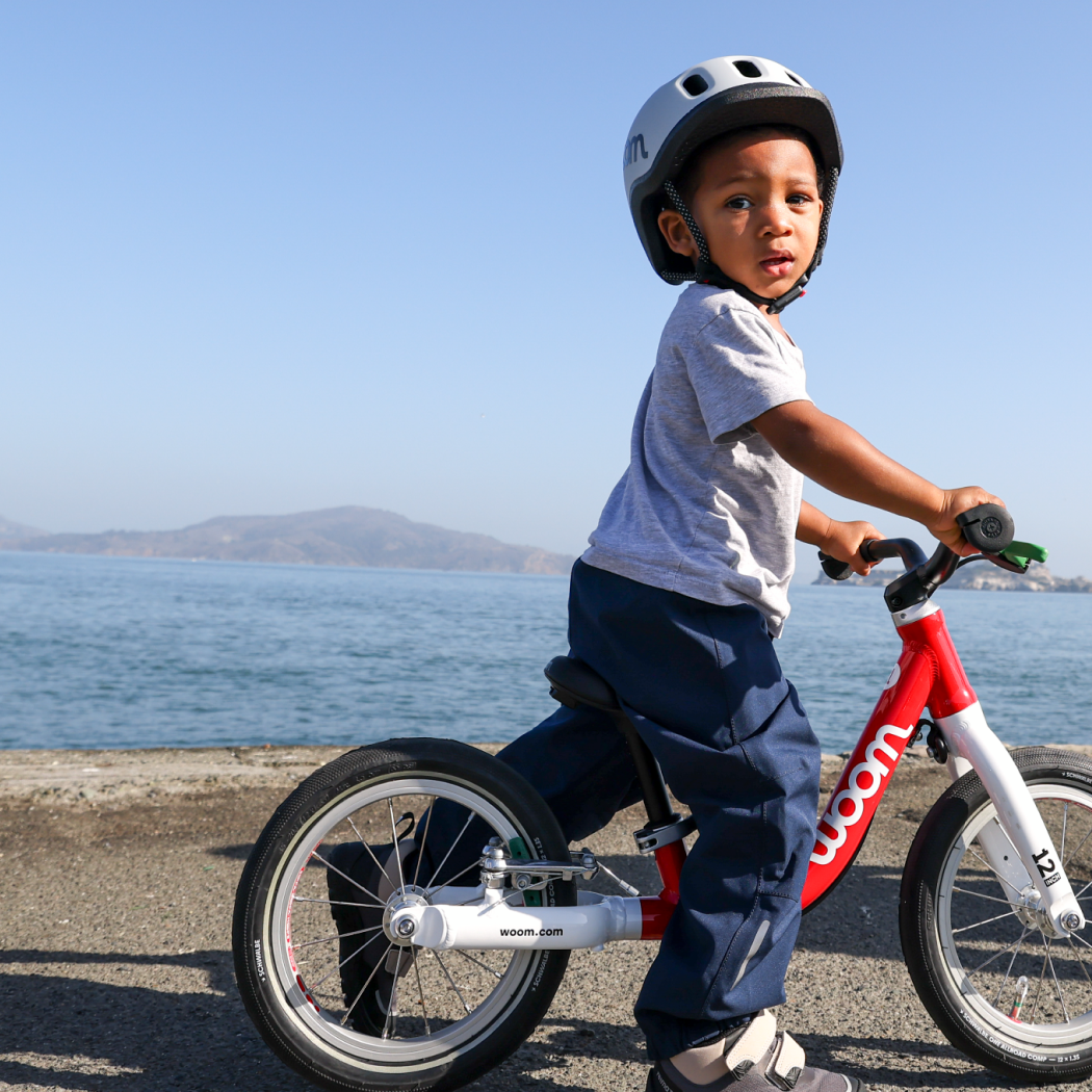 A boy wearing a woom grey READY helmet, riding a red woom GO bike by the San Francisco bay.