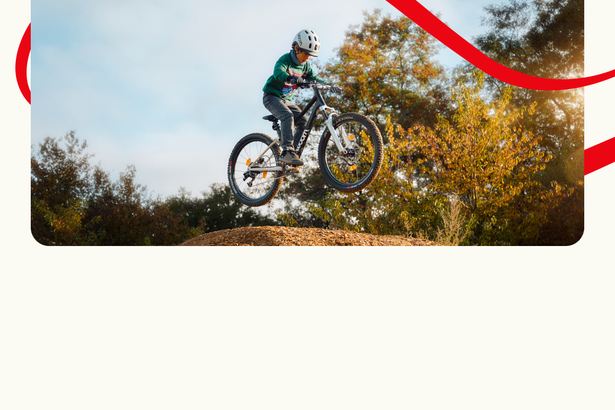 Ein Kind auf einem Woom OFF Mountainbike springt hoch über eine Erdrampe, im Hintergrund sind Bäume und der Himmel zu sehen. Dekorative rote Linien umrahmen die Szene.