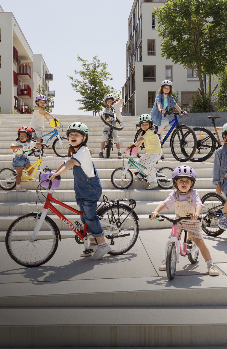 Happy kids on woom bikes wearing woom READY helmets, gathered on wide outdoor steps in a modern residential area.