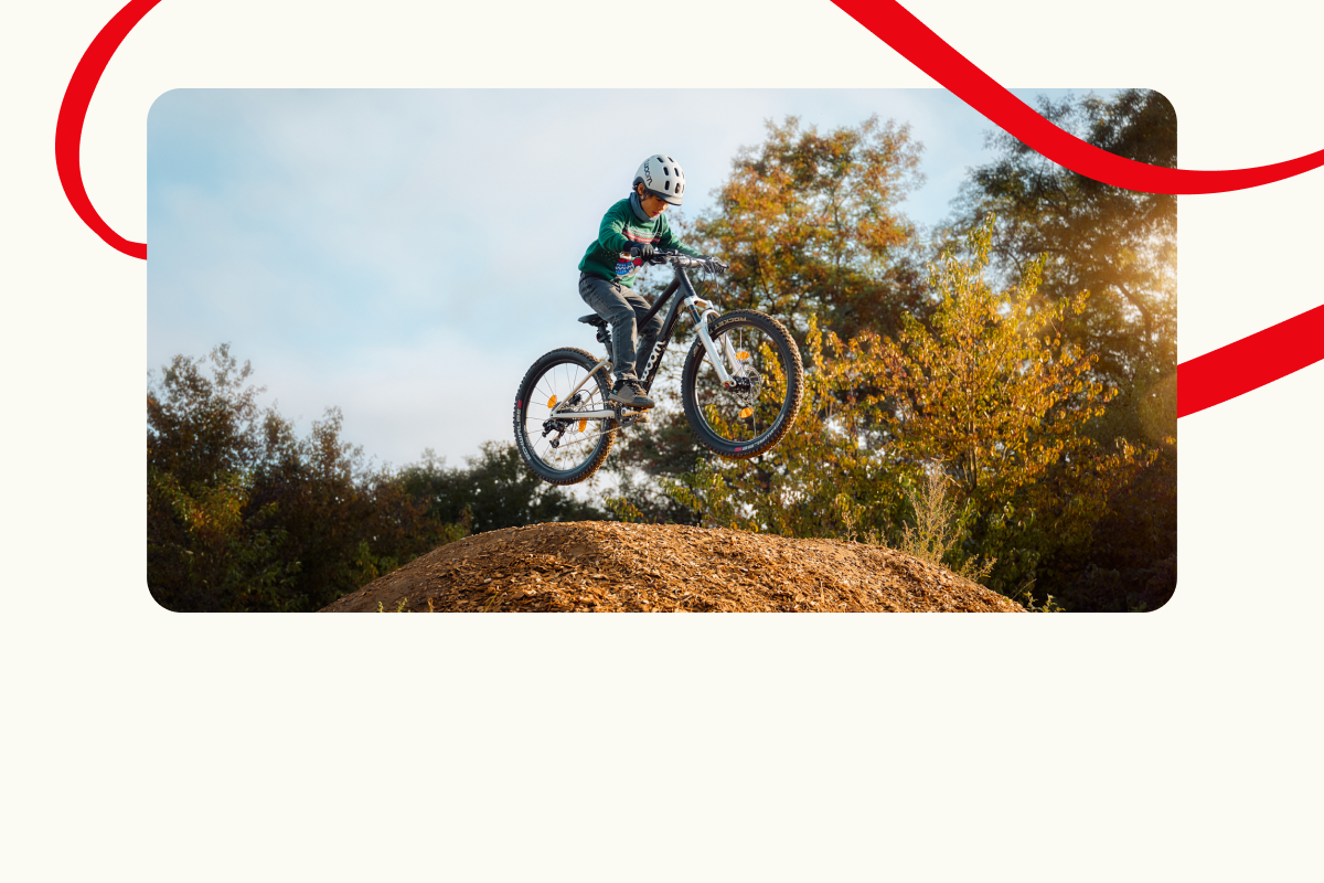 A child on a woom OFF mountain bike is jumping high over a dirt ramp with trees and sky in the background. Decorative red lines frame the scene.