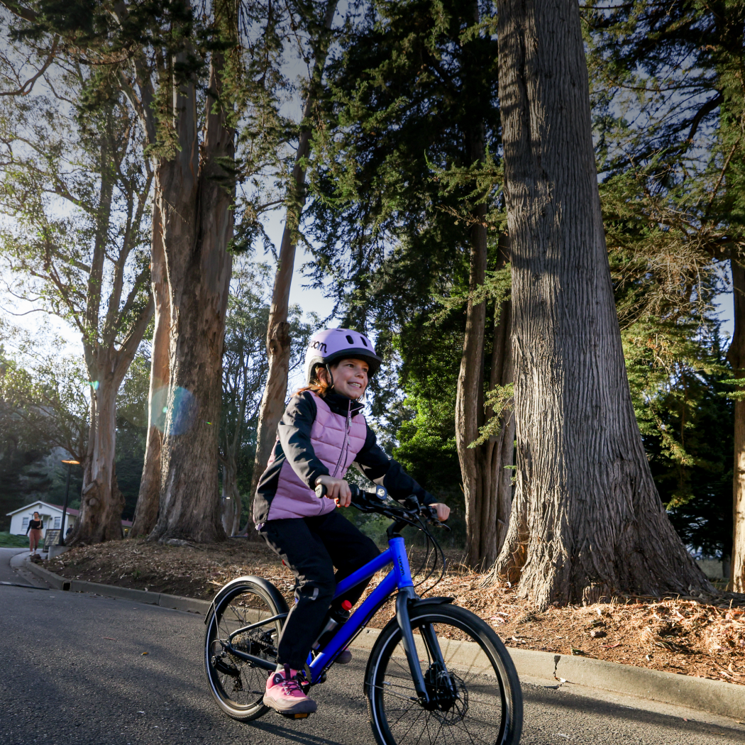 A girl in a warm pink jacket rides her magnetic blue EXPLORE bike while wearing her READY Helmet through a neighborhood street, with the sun low in the sky behind her.
