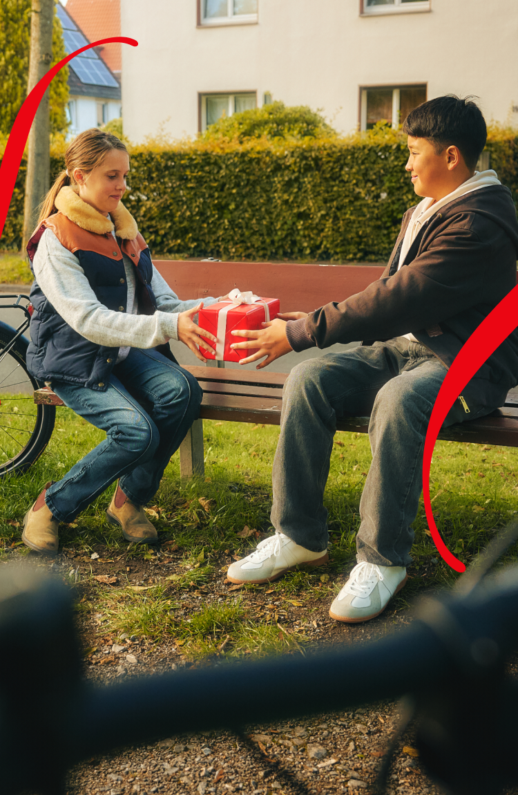 A boy and a girl are sitting on a park bench, and the boy is handing the girl a red-wrapped gift.