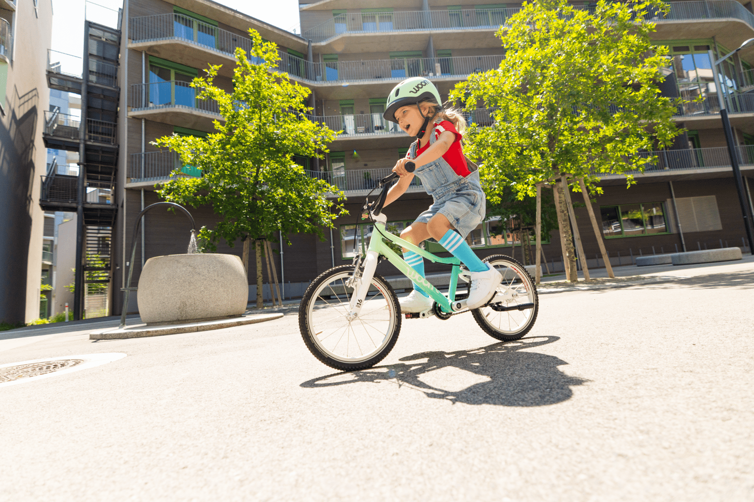 A girl wearing a mint-colored READY helmet rides past an apartment building on a spearmint crush woom GO.