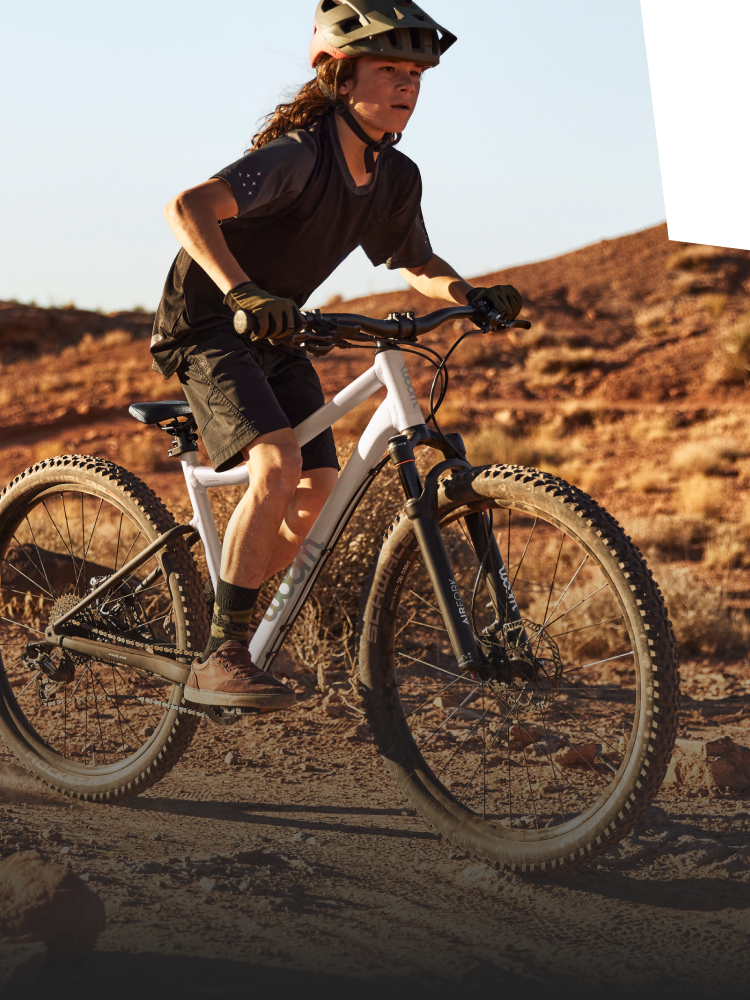 Junge mit Fahrradhelm auf dem Kopf fährt auf einem weißen Mountainbike mit Federgabel von woom auf einem ruppigen Trail.