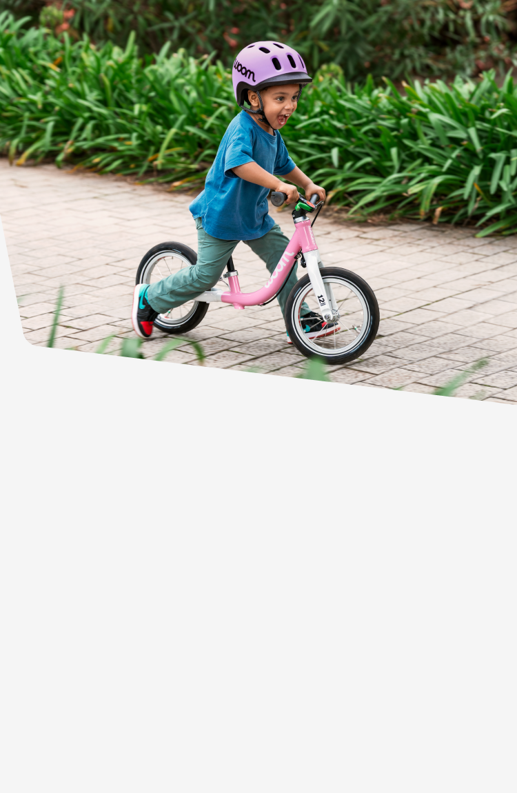 A young boy grins while riding a powder pink woom GO 1 down a brick path lined with bushes wearing a lilac READY Helmet