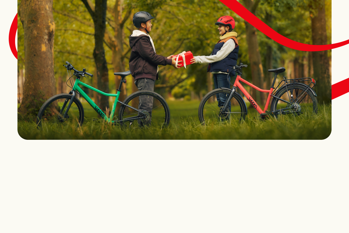 Zwei Kinder mit woom EXPLORE-Fahrrädern tauschen in einem Wald ein Geschenk aus. Dekorative rote Linien umrahmen die Szene.