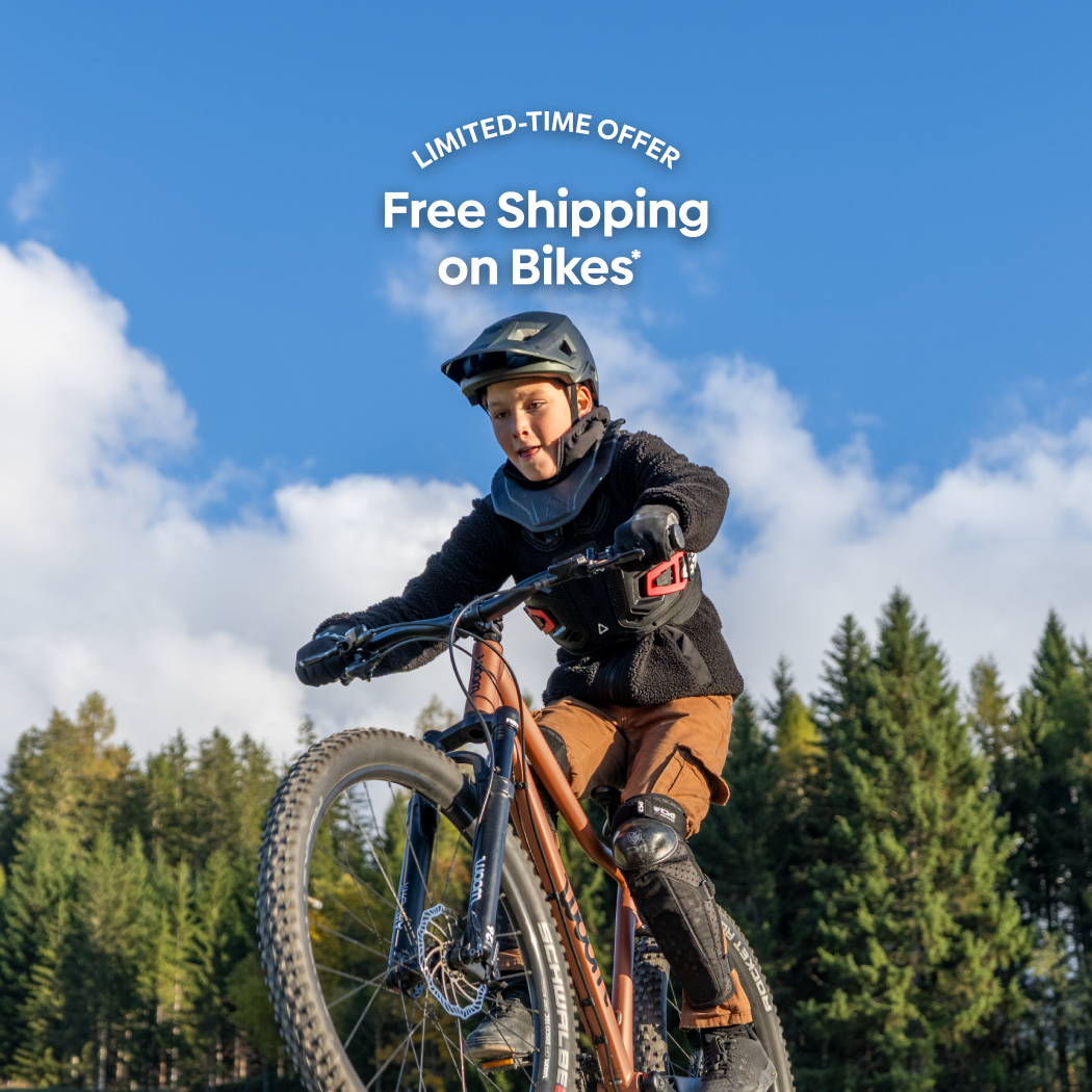 A boy in winter wear and a helmet fiercely concentrates while popping a wheelie on a terra coppa-colored woom OFF AIR mountain bike in a tree-lined lush green field.