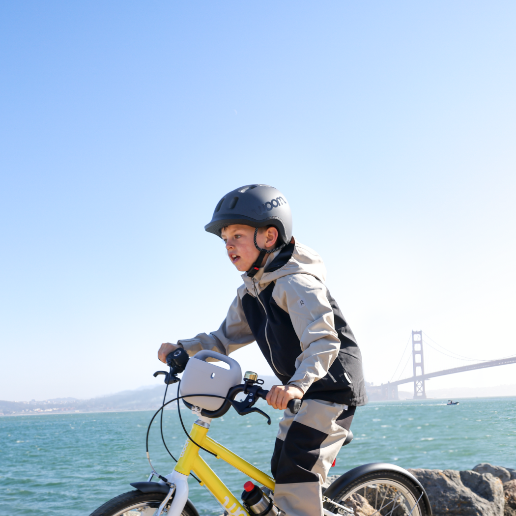 A boy rides a vibrant yellow woom GO bike, complete with a POP Basket attached, on a waterside trail while wearing a READY Kids’ Helmet with the Golden Gate Bridge visible in the background