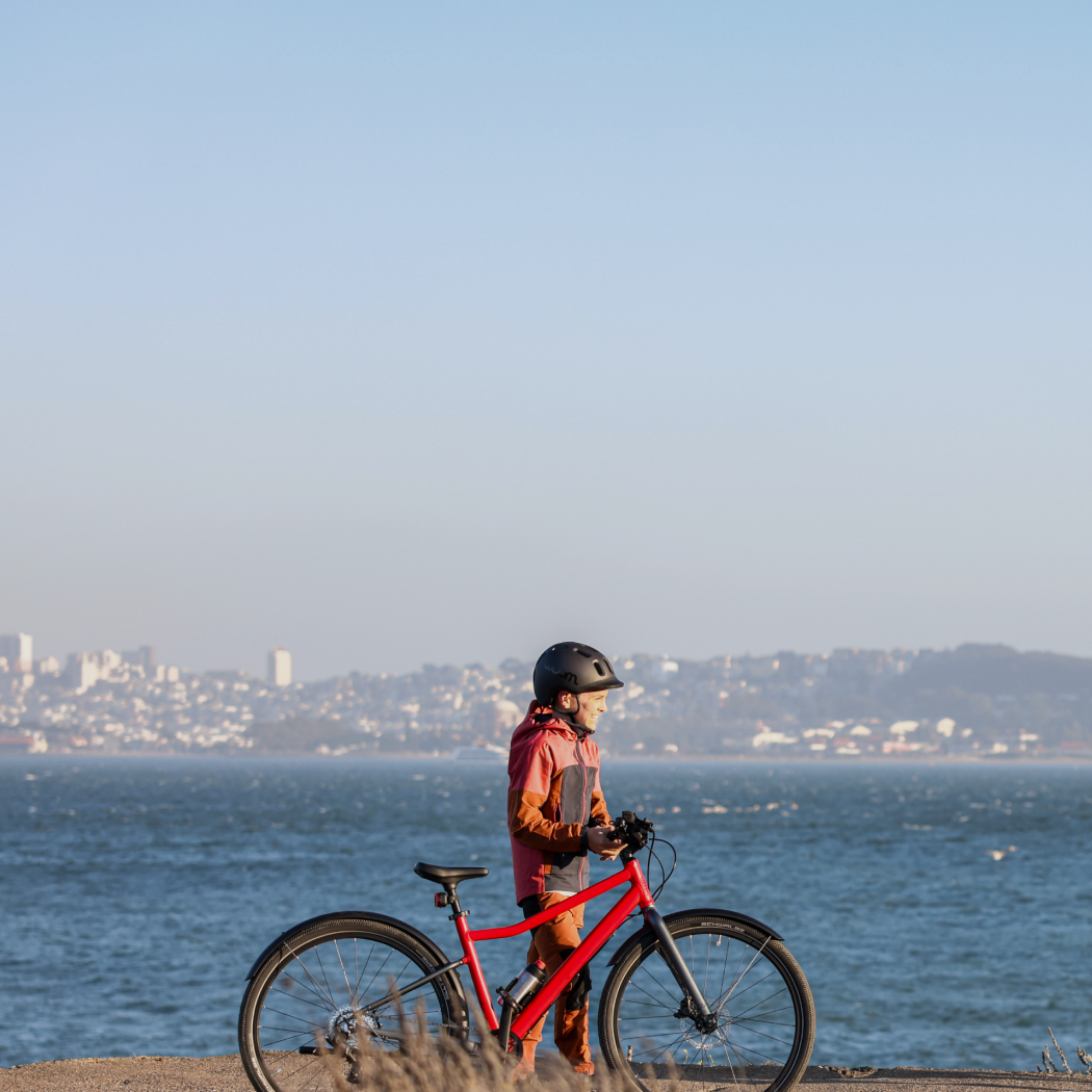 A boy wearing a READY Kids’ Helmet stands next to his woom red EXPLORE bike while looking out at the San Francisco Bay, accompanied by his dog