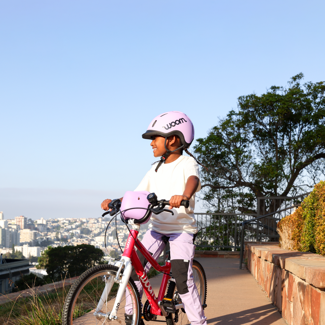 A young girl in braids and a lilac READY Kids’ Helmet sits atop her hot pink woom GO 4 bike, equipped with POP Bike Basket and BEAM Bike Light, overlooking a neighborhood from the top of a tall hill at golden hour.