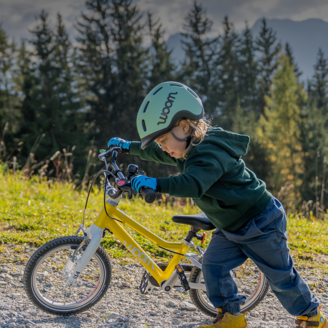 A young child in a mint READY Kids’ Helmet and sky blue TENS Bike Gloves focuses intensely on pushing his vibrant yellow woom GO 2 up a tree-lined incline.