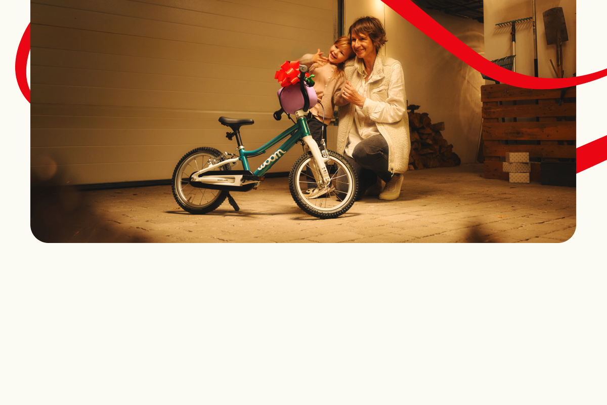 Ein Erwachsener kniet neben einem Kind und einem Woom GO vor einer Garage. Dekorative rote Linien umrahmen die Szene.
