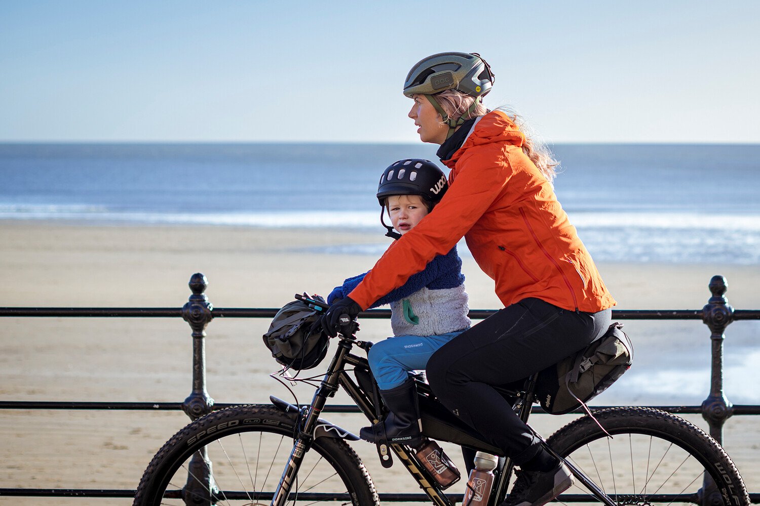 Eine Mutter radelt am Meer mit ihrem Sohn, der im Front-Kindersitz sitzt und einen schwarzen Fahrradhelm von woom trägt.