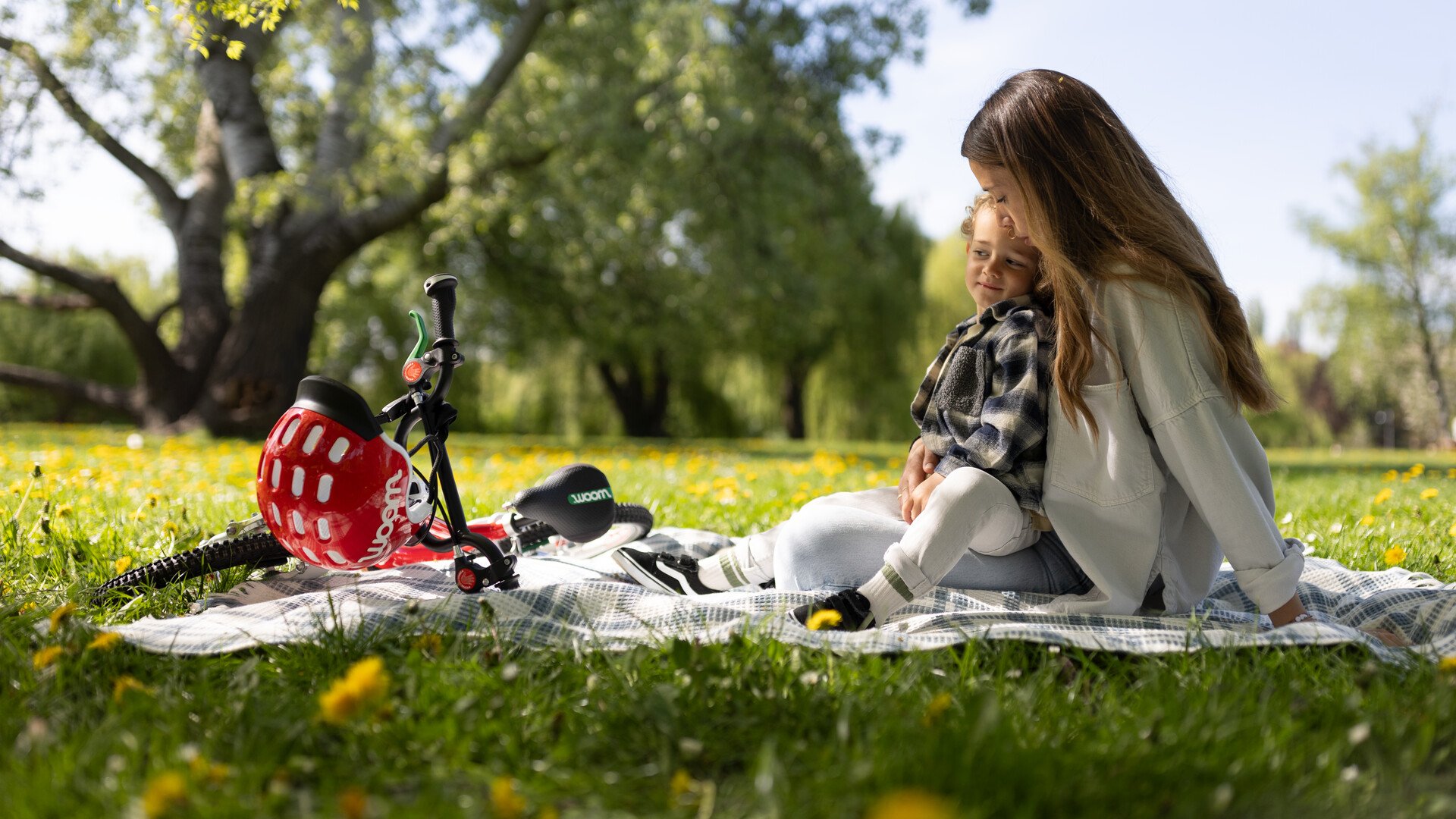 Ein kleiner Junge und seine Mutter sitzen auf einer Picknickdecke im Park neben einem woom Kinderfahrrad.