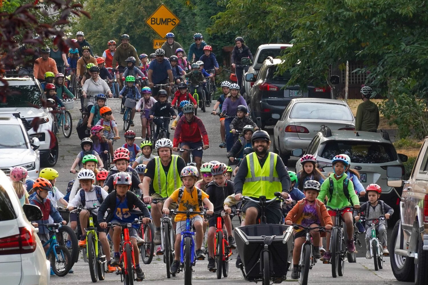 Coach Sam Balto leads a Bike Bus ride down a car-lined street with dozens of kids behind him. Image credit: Sam Balto/Bike Bus World