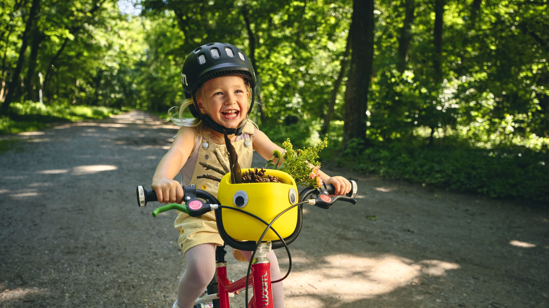 Ein kleines, lachendes Mädchen mit schwarzem woom Helm fährt auf einem rot-weißen woom bike auf einem schattigen Waldweg. Auf dem Lenker steckt ein gelber Fahrradkorb fürs Kinderrad.