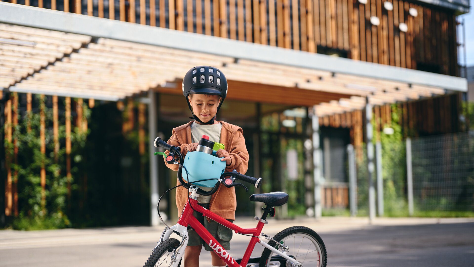 Ein kleiner Junge steckt den hellblauen woom Fahrradkorb fürs Kinderrad vorne auf den Lenker seines rot-weißen woom bikes.