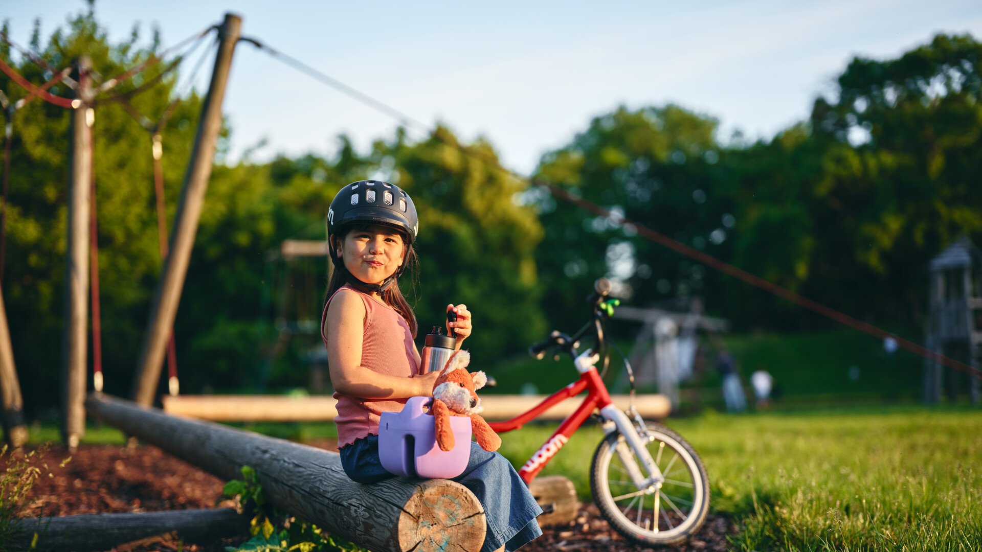 Ein kleines Mädchen mit schwarzen Haaren, dunklen Augen und schwarzem woom Helm sitzt auf einem Spielplatz inmitten einer Wiese. Neben ihr steht ein lilafarbener woom fahrradkorb in dem ein Fuchs-Kuscheltier steckt. In der Hand hält sie die Trinkflasche von woom.