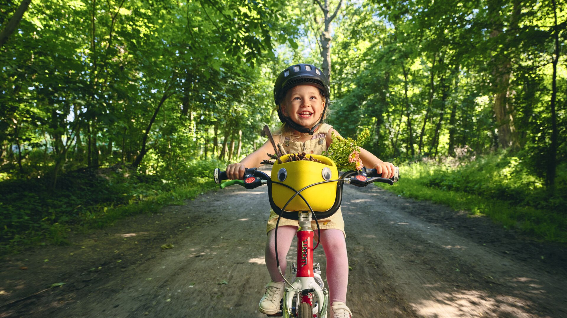 Ein kleines, lachendes Mädchen mit schwarzem woom Helm fährt auf einem rot-weißen woom bike auf einem schattigen Waldweg. Auf dem Lenker steckt ein gelber Fahrradkorb voll mit kleinen Schätzen aus der Natur.