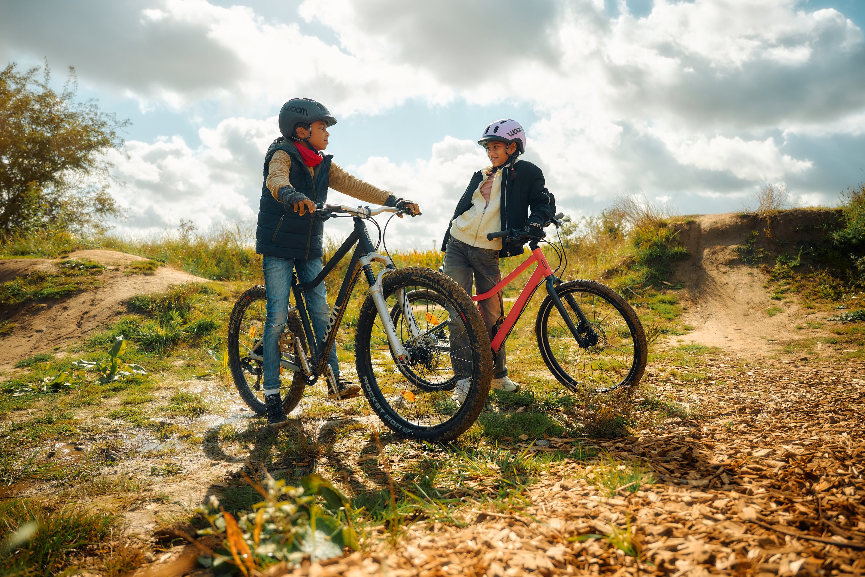 Two children with helmets on mountain bikes (woom OFF and woom EXPLORE) on a dirt track.