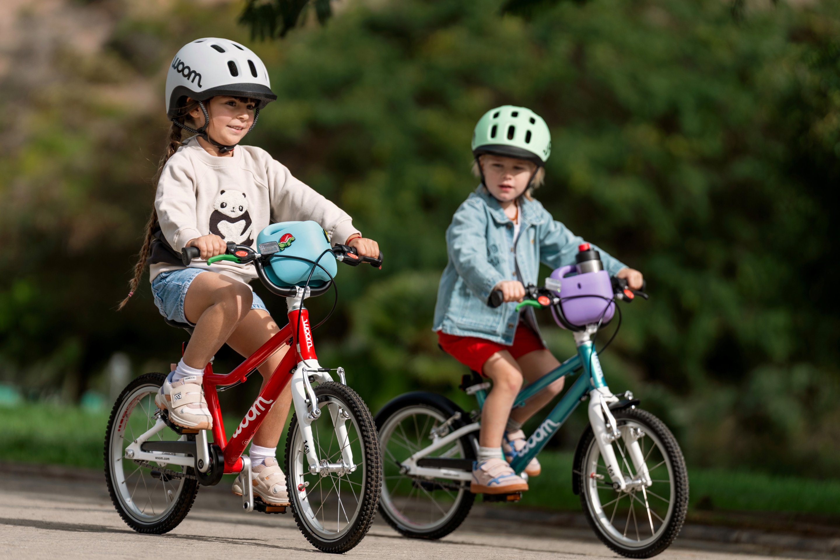 Zwei Kinder auf woom GO Bikes mit woom READY Helmen und bunten POP Fahrradkörben fahren nebeneinander auf einem Weg.