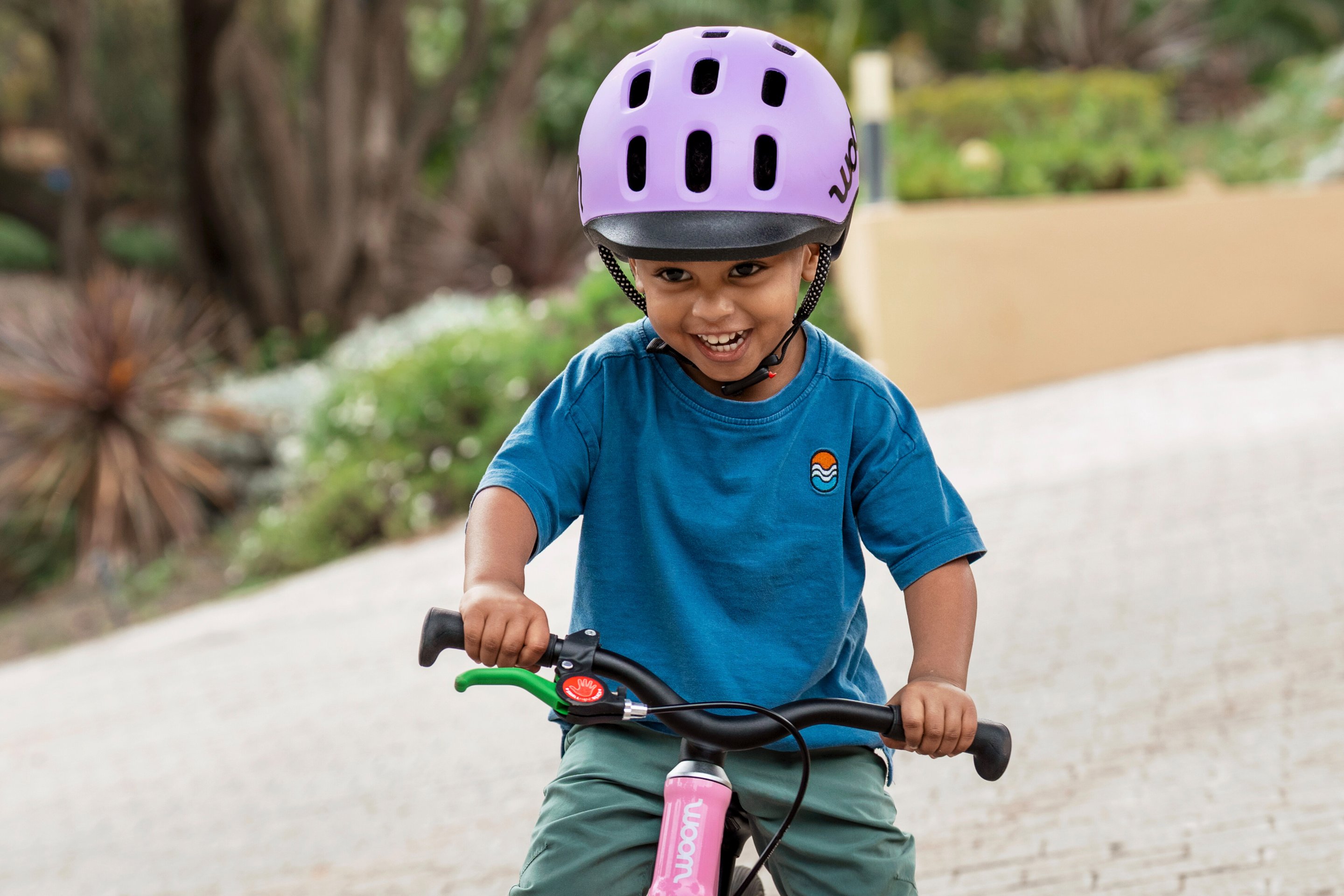 Un enfant souriant portant un casque READY violet roule avec sa draisienne woom GO rose.