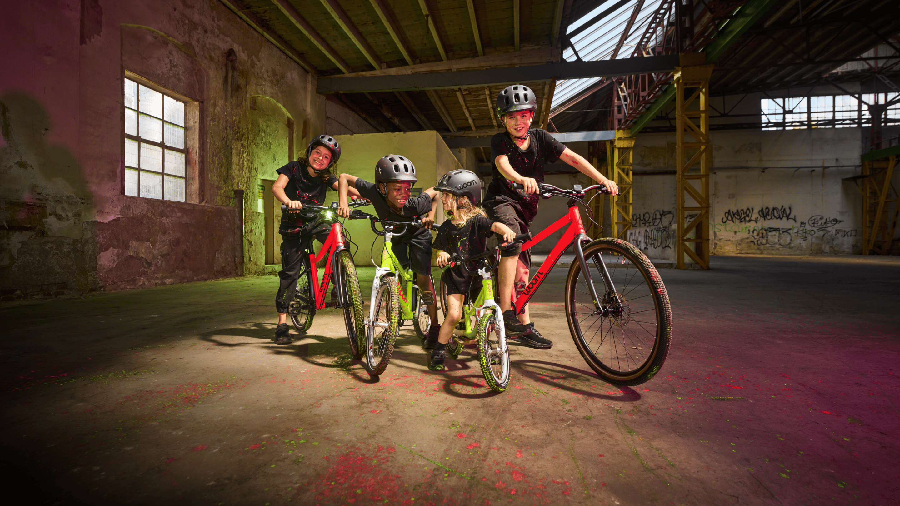 Vier Kinder mit woom Helmen posieren mit leuchtend neonpinken und limettengrünen woom Bikes in einer rustikalen Industriehalle.