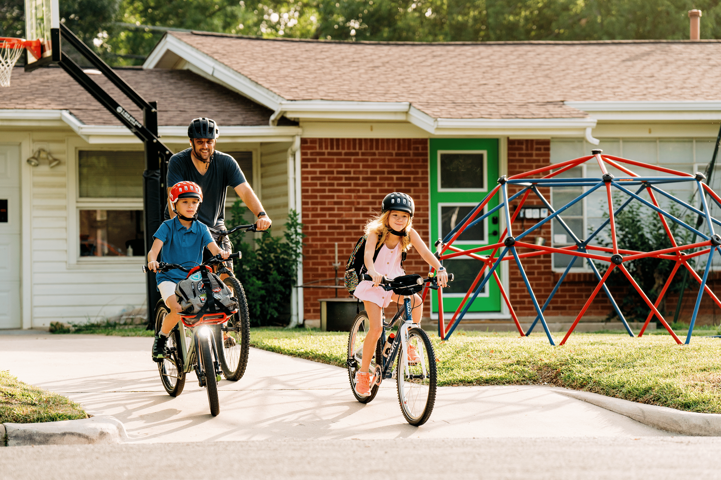 A father and his two kids, backpacks strapped on, ride their woom bikes out of their driveway to begin their journey to school.