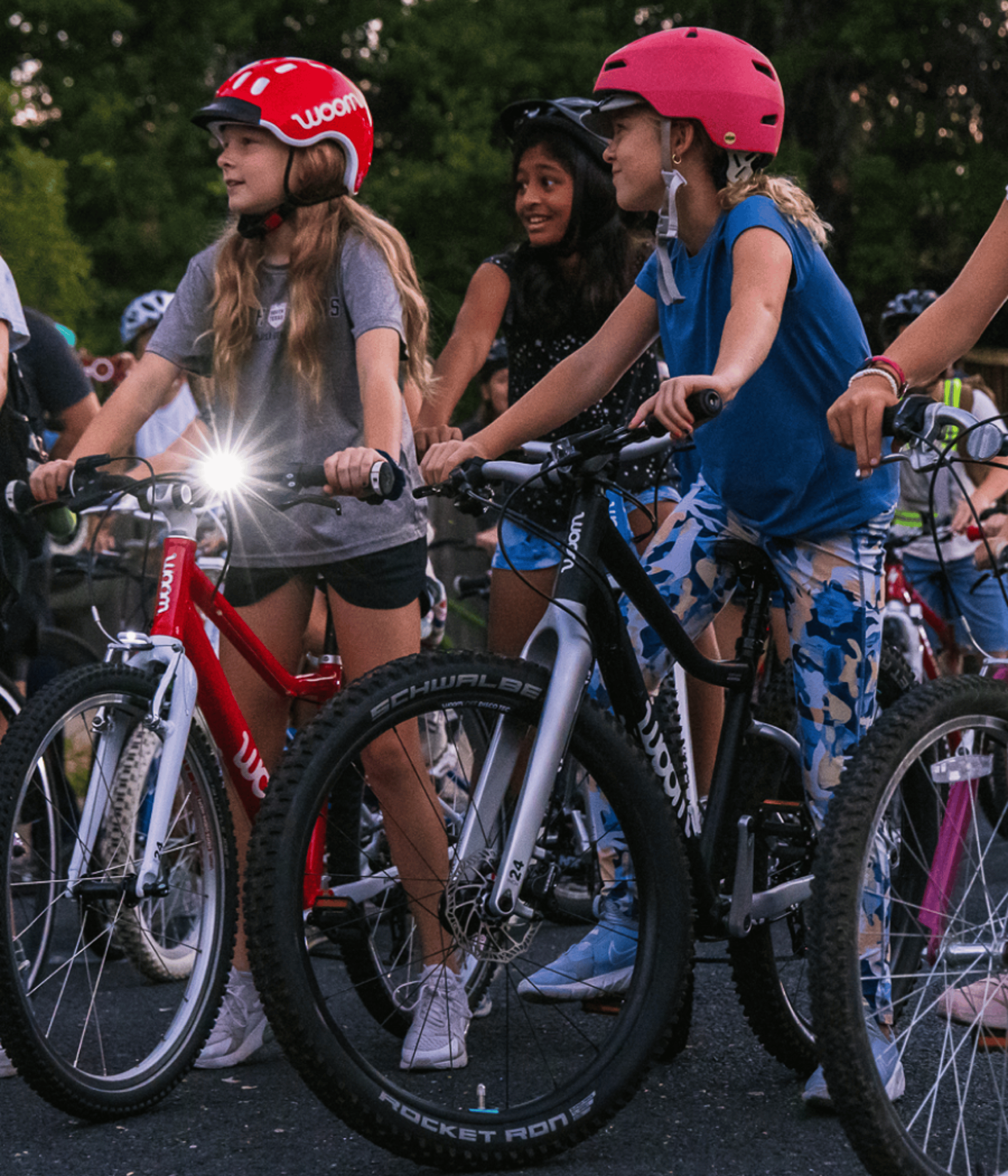 Two preteen girls wait on their woom bikes for the Bike Bus ride to begin, with one girl’s bike light flashing in the dusky light.