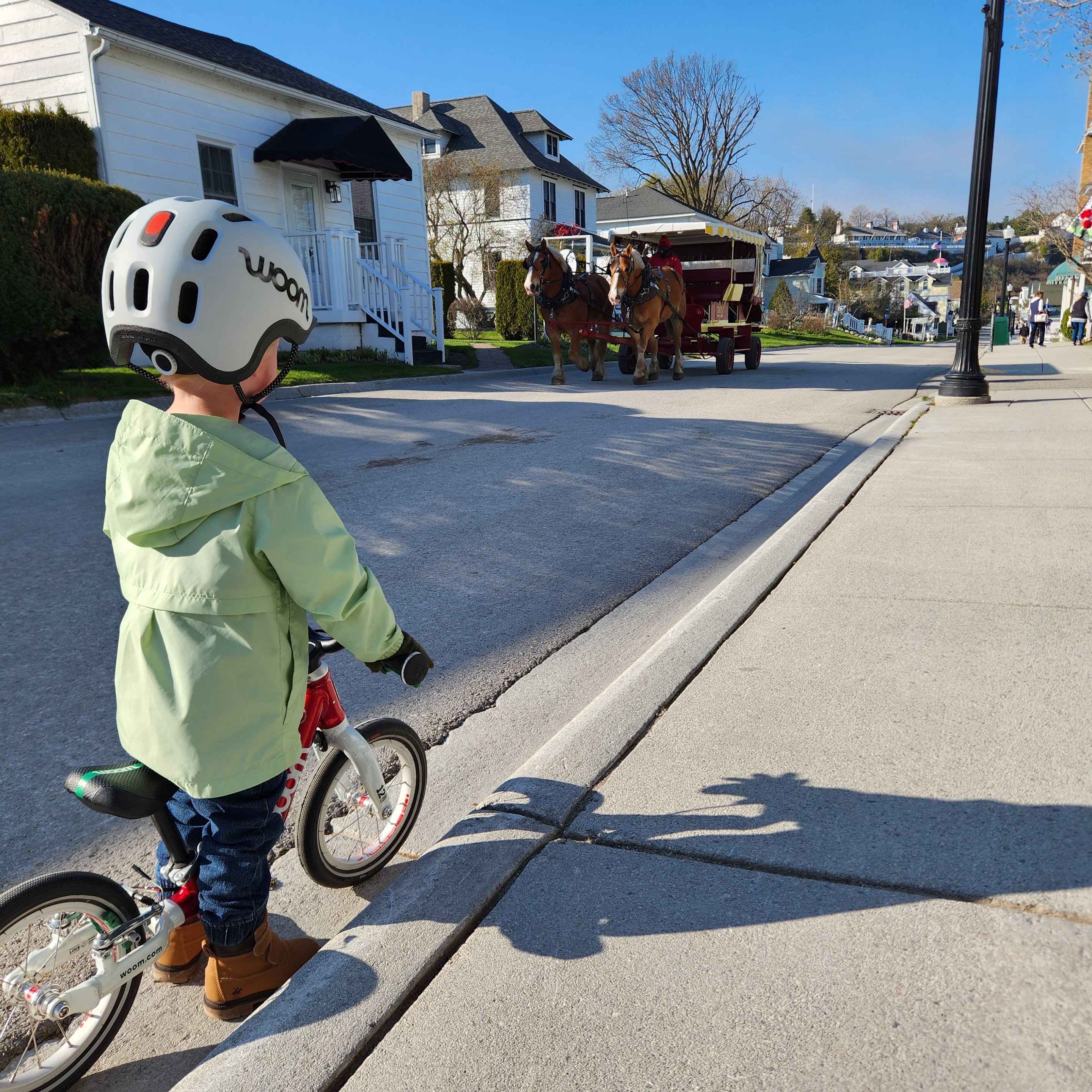 A 3-year-old boy faces away from the camera in the left of the image wearing a light grey READY Kids’ Helmet with READY Helmet and a mint green raincoat. He is straddling his red woom ORIGINAL 1 bike and watching clydesdales pull a horse-drawn carriage down an empty neighborhood street.