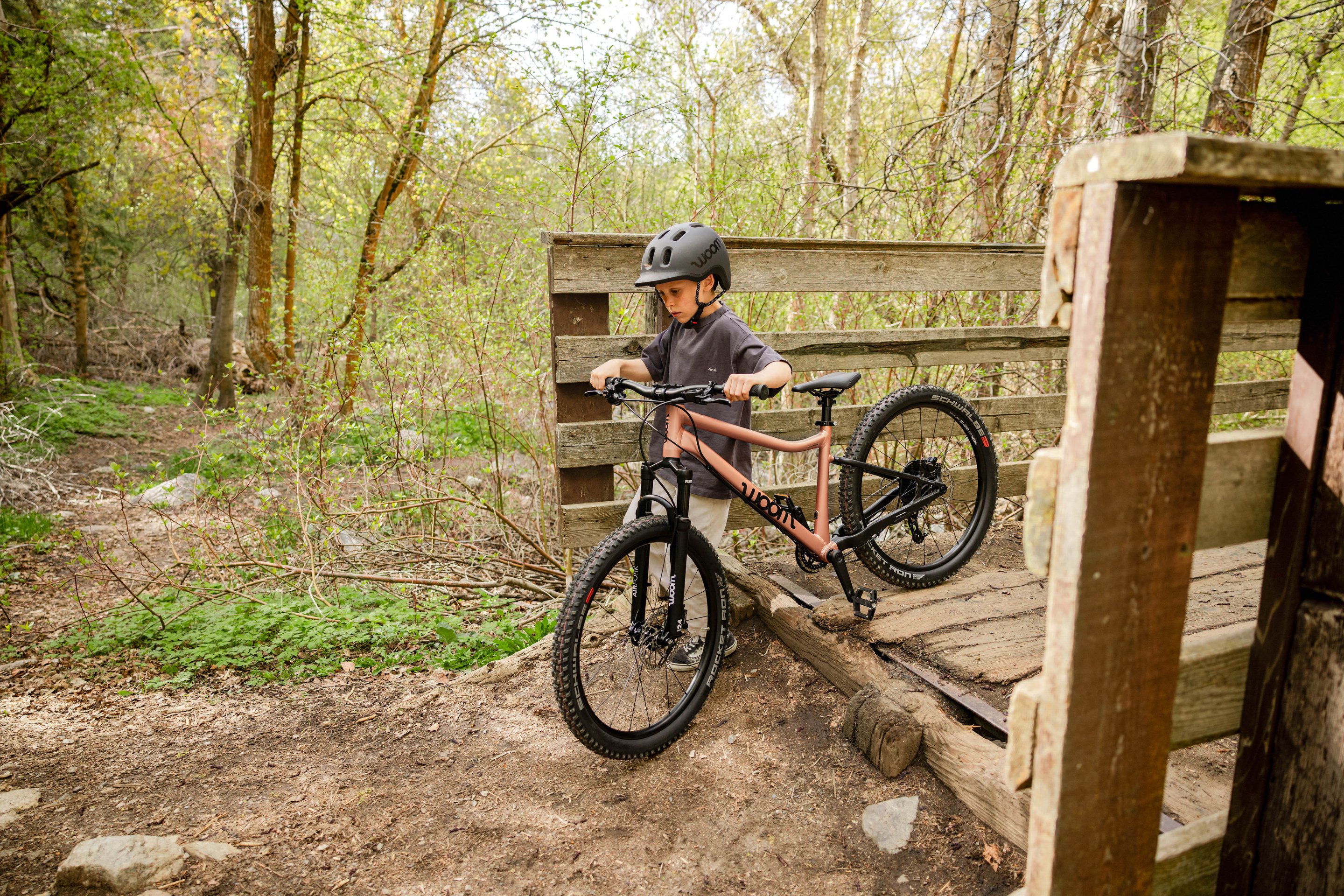 A photo in a sunlit forest of a young boy walking his woom OFF AIR bicycle off of a wooden bridge.