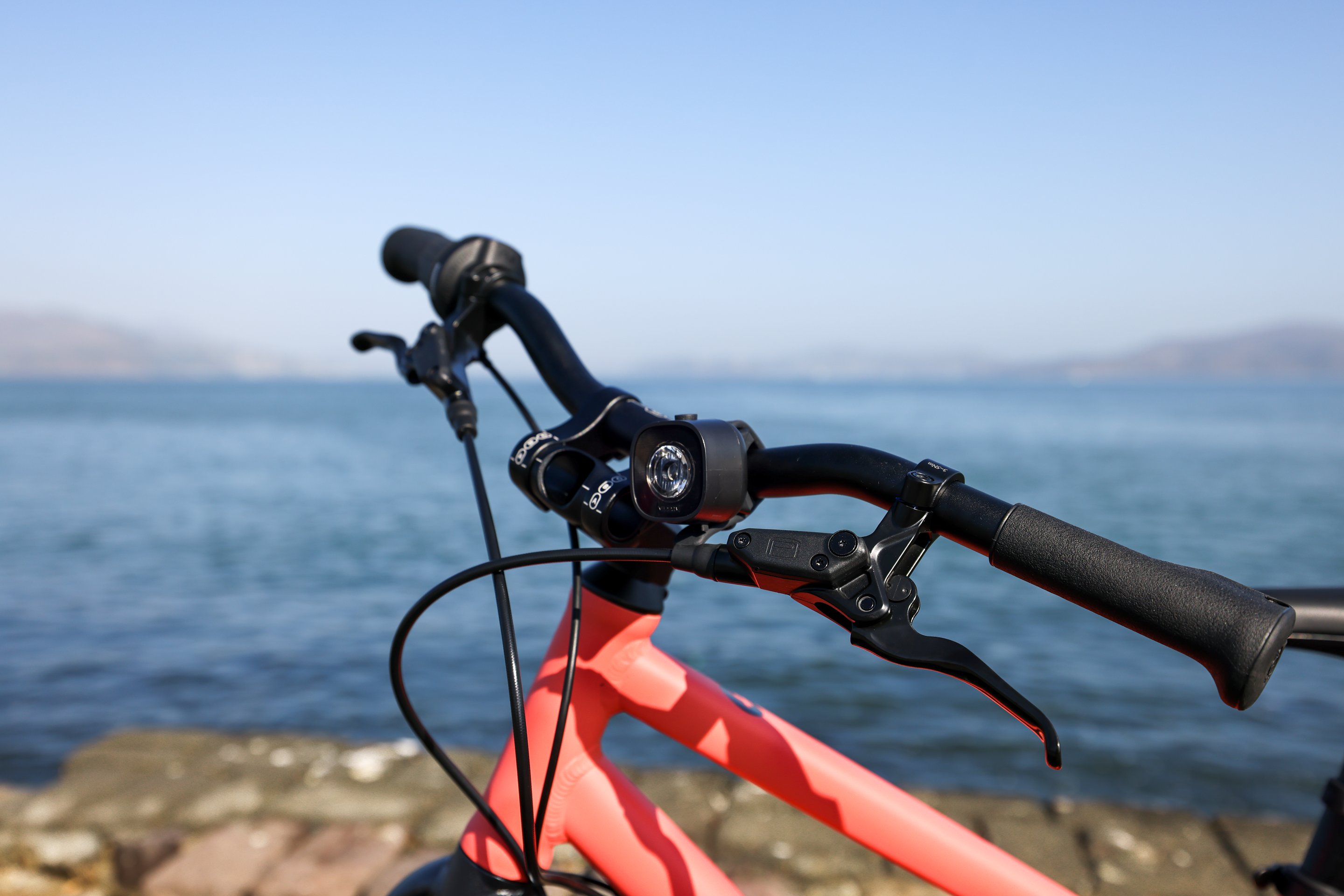 A close-up photo of the handlebars, stem, and frame of a neon coral woom EXPLORE bike, complete with BEAM light, against a bayside backdrop.