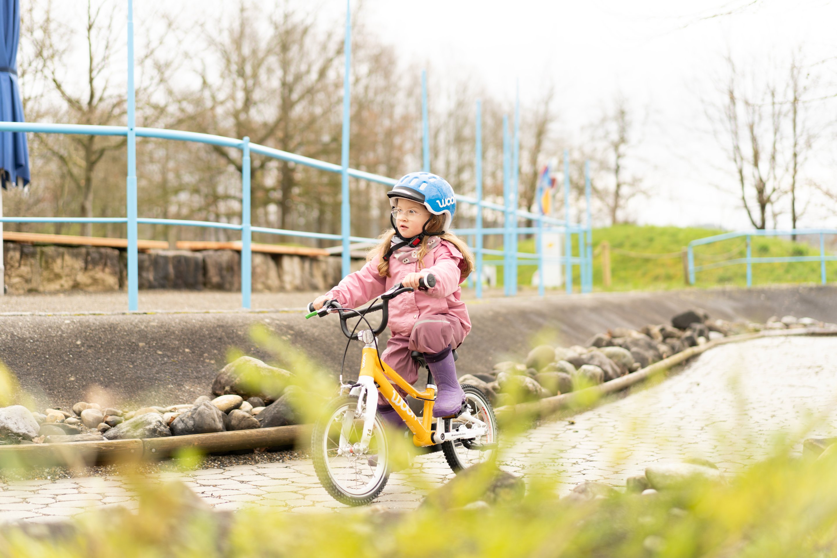 A young girl rides a woom ORIGINAL through a park on a grey day.