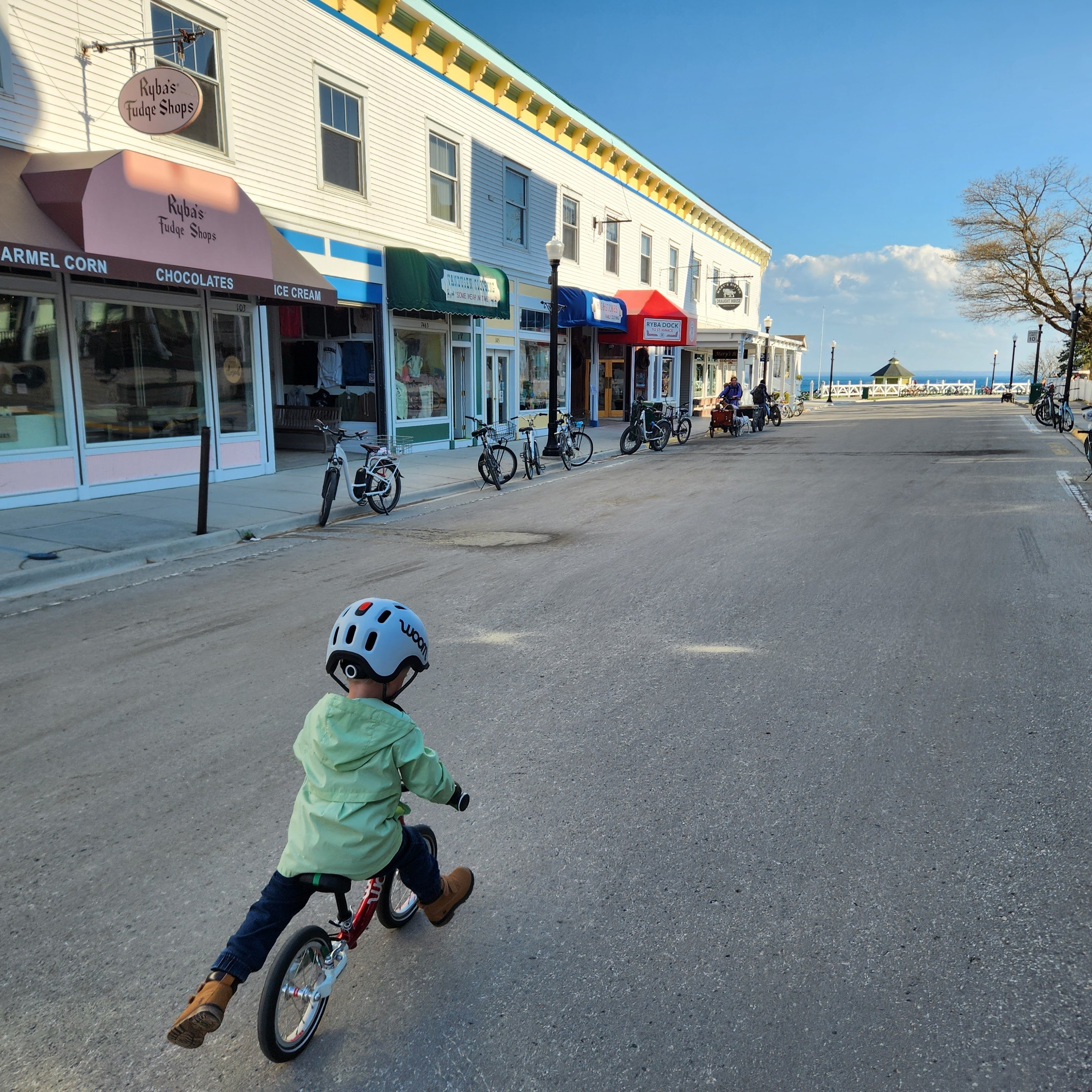A three-year-old boy in a mint green raincoat, boots, jeans, and light grey READY Kids’ Helmet with READY Helmet Light confidently rides his woom ORIGINAL 1 balance bike into frame, away from the camera, down a quaint store-lined street with Lake Michigan visible in the background.