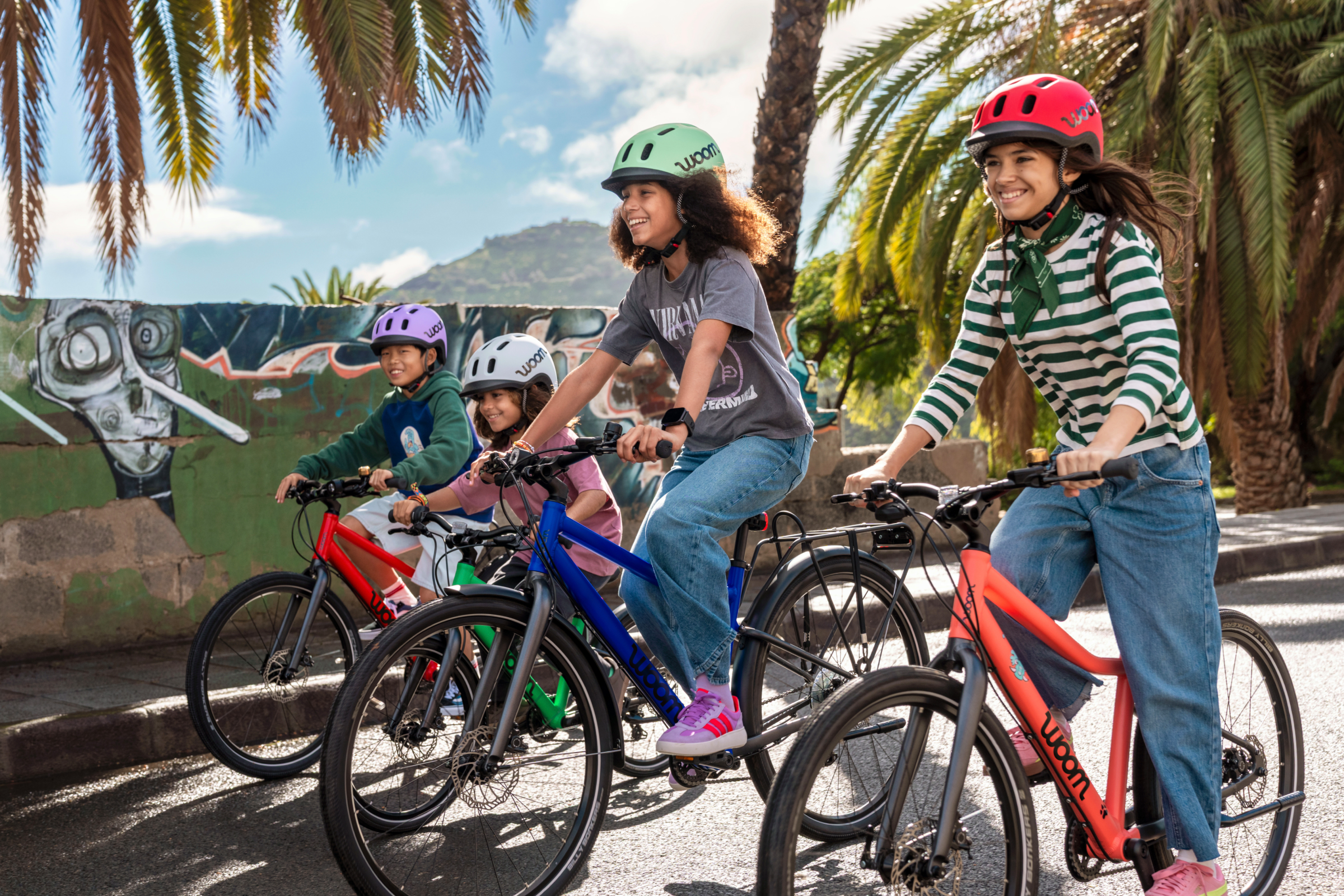 Four children riding woom EXPLORE bikes wearing woom READY helmets on a sunny street with palm trees.