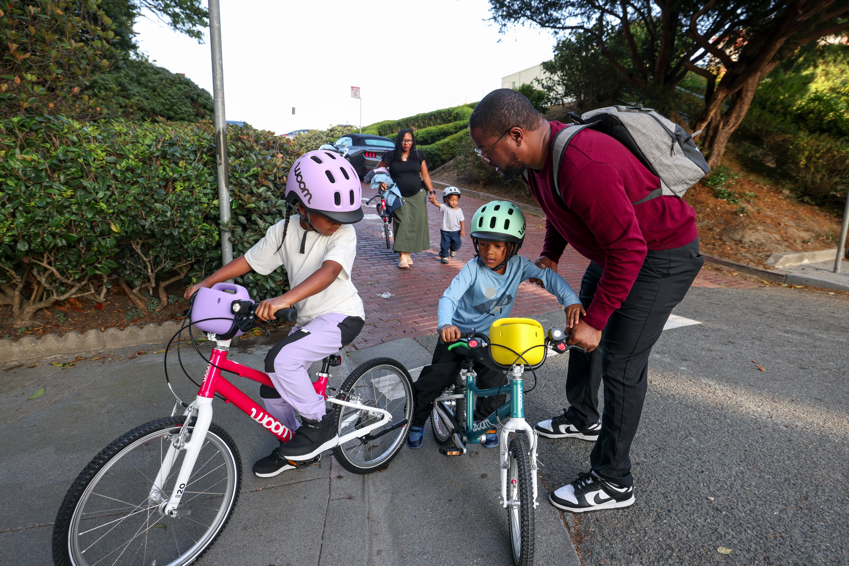 A photo of a dad preparing his two kids on their GO bikes for a ride, with mom and toddler holding hands in the background as she carries his small woom GO 1 bike.