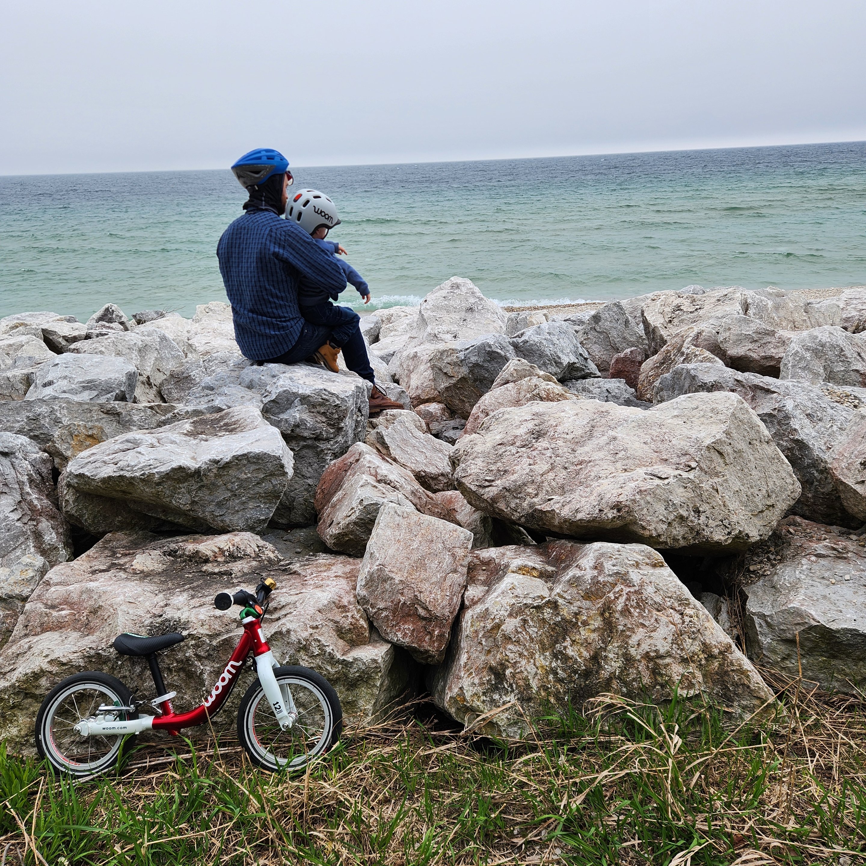 A father in a blue shirt, jeans, boots, and a blue helmet holds his son on his lap while they sit on rocks along the lakeshore and watch the waves. The boy is wearing a light grey READY Kids’ Helmet with READY Helmet Light and his red woom ORIGINAL 1 bike is leaned agains the rocks near the bottom left of the image.