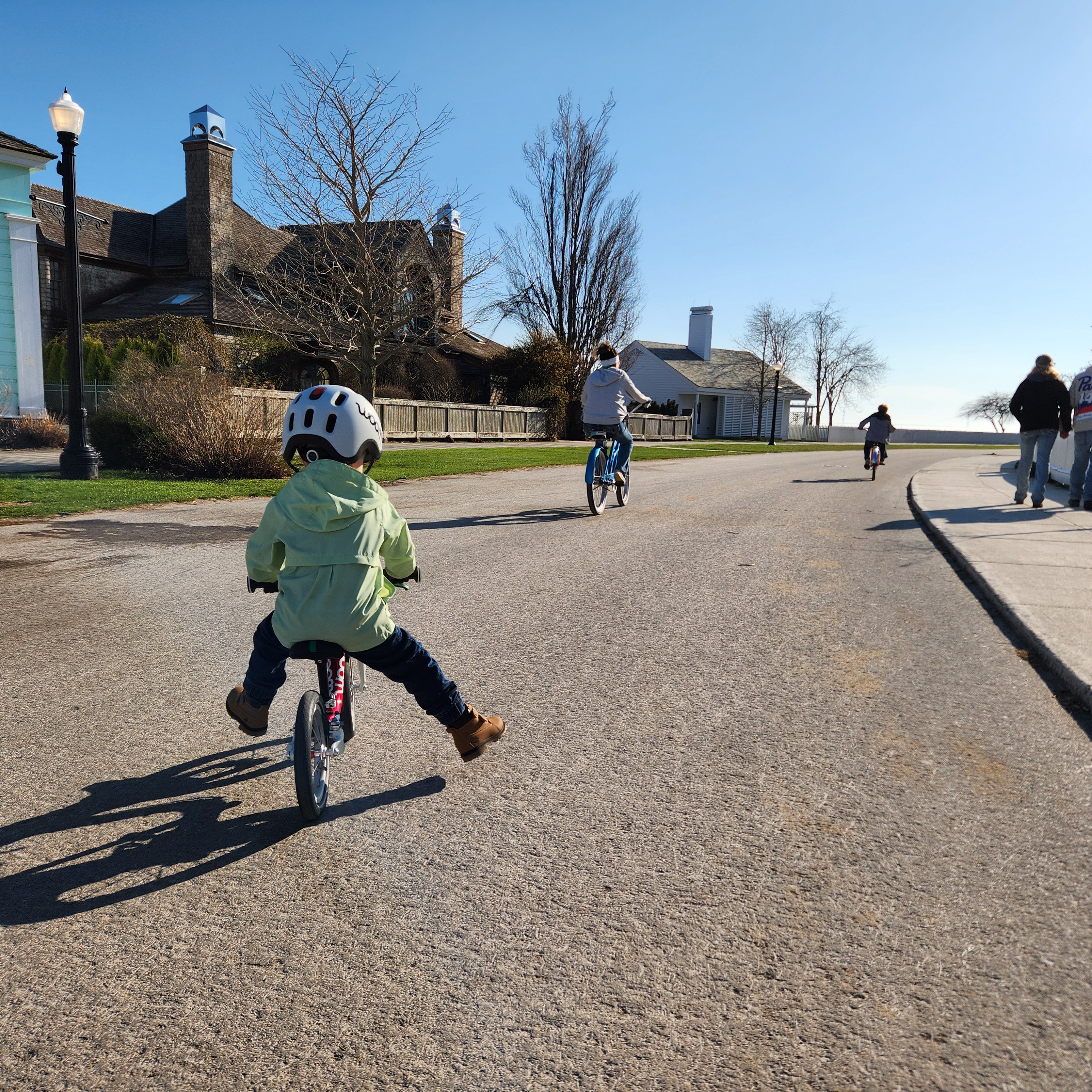 A 3-year-old boy with his back to the photographer wearing a light grey READY Kids’ Helmet with READY Helmet Light attached. He is riding a red woom ORIGINAL 1 balance bike on a neighborhood road with two other cyclists on a cloudless day.