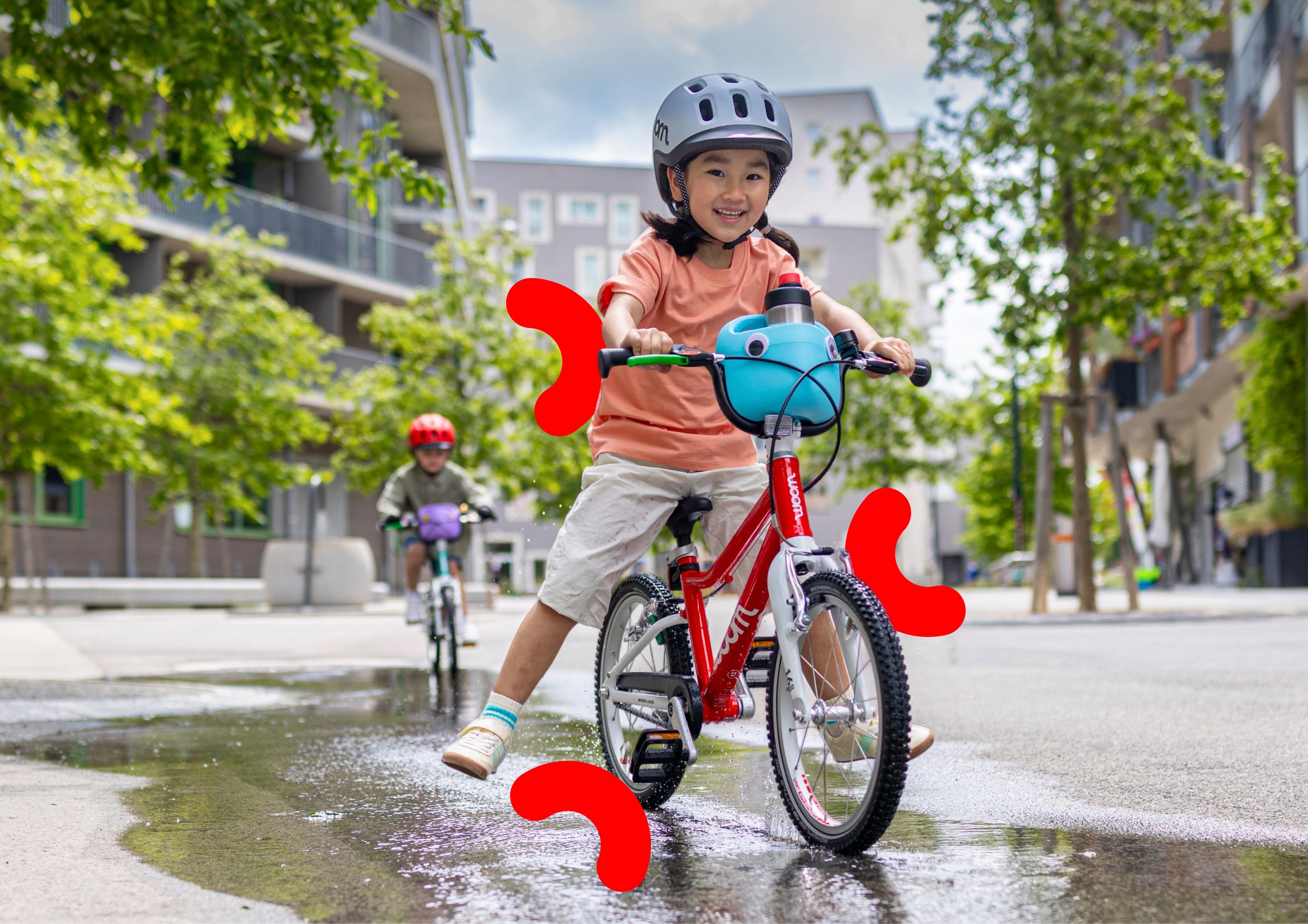 Child in a grey woom READY helmet riding a woom red woom GO bike through a puddle in an urban setting with a second rider behind.
