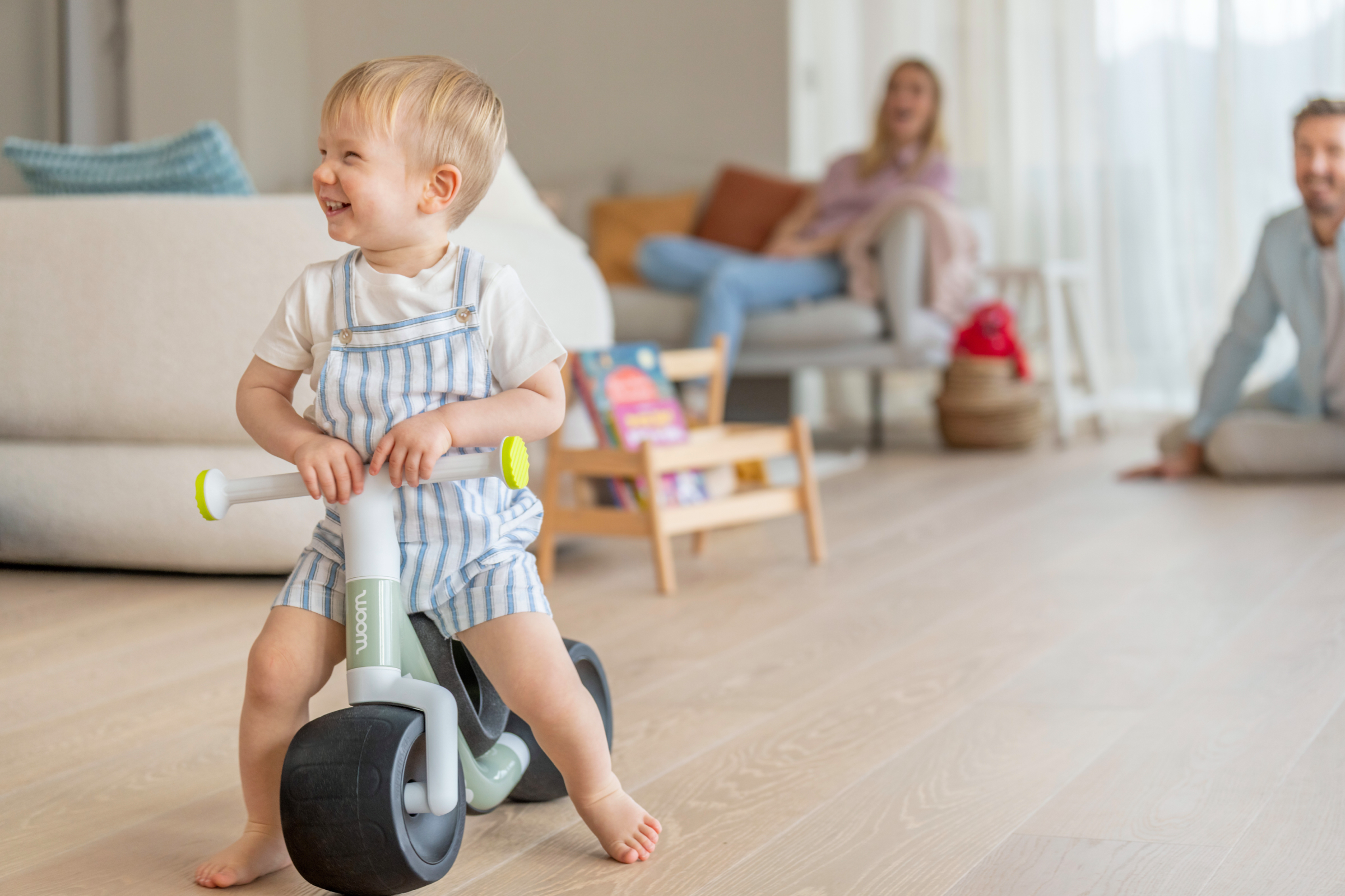 Laughing toddler riding a soft sprout woom WOW balance bike in a bright living room.