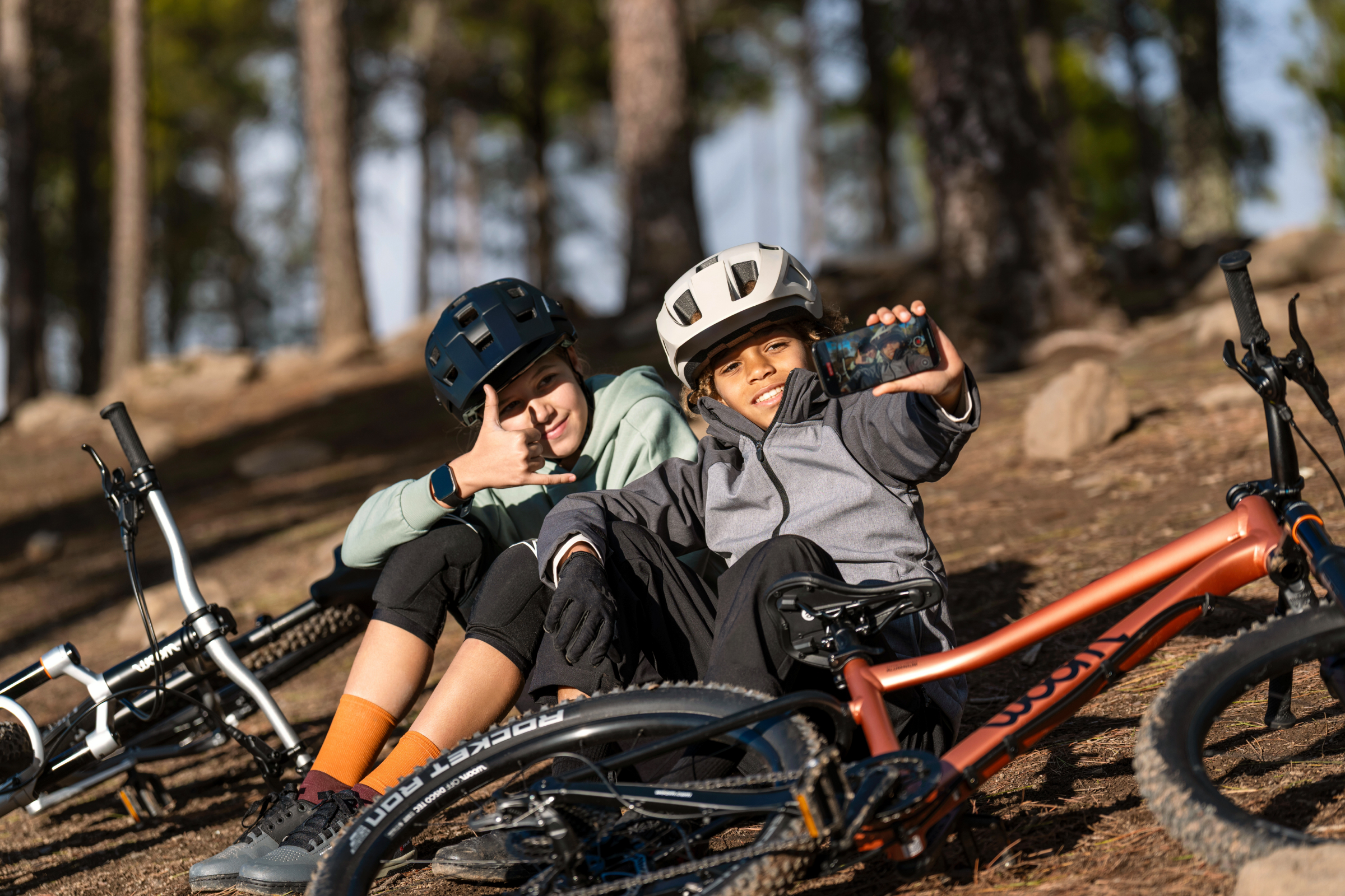 Two children taking a selfie next to their woom OFF mountain bikes in a sunny forest area.