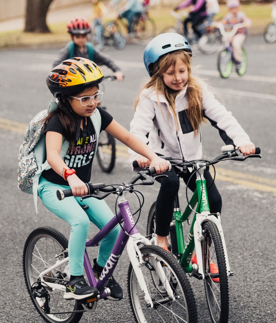Two girls on green and purple woom bikes focus on their pedaling, with kids following behind them down the street.