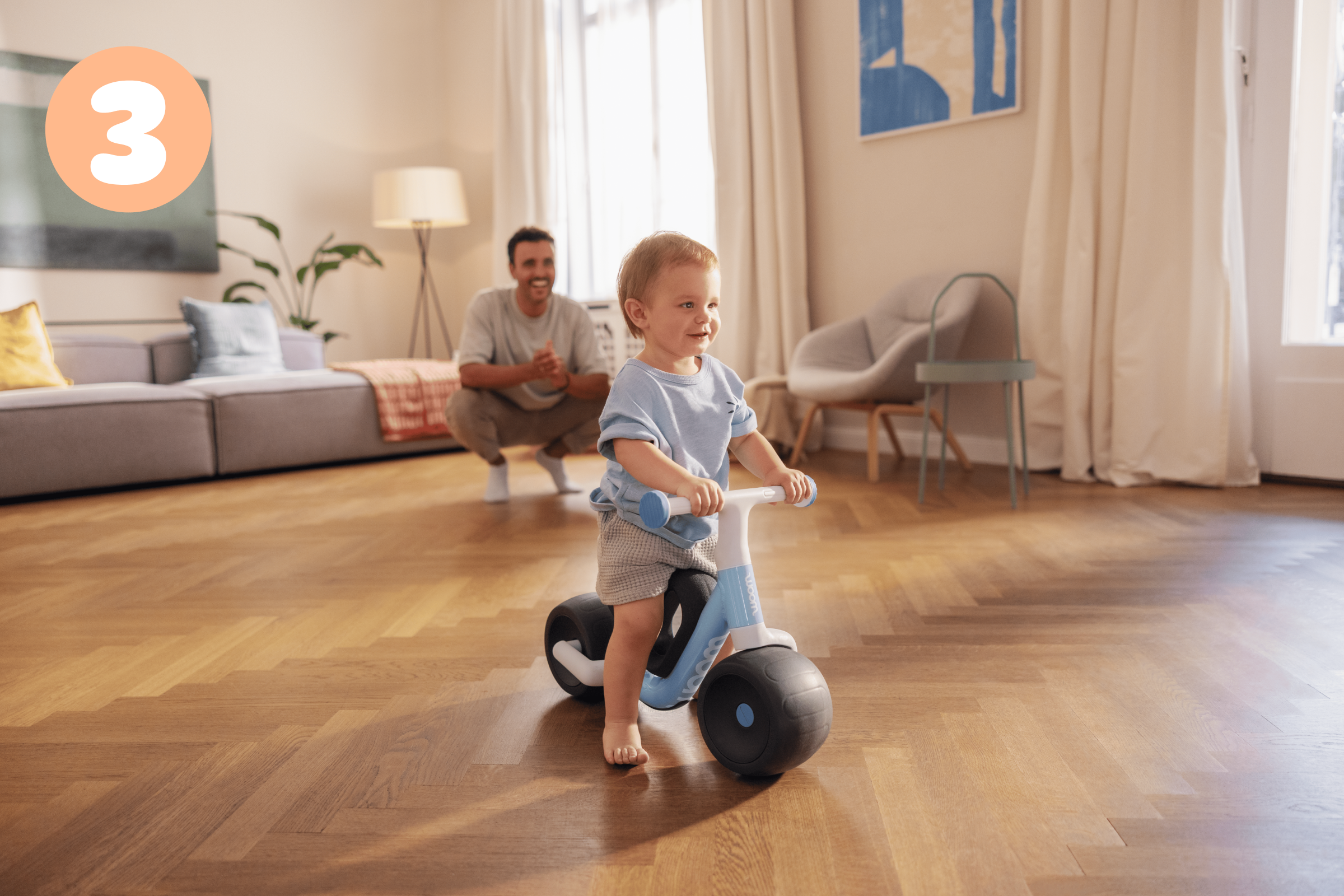A toddler is sitting on a light blue woom WOW bike in a living room, with a man kneeling on the floor in the background.