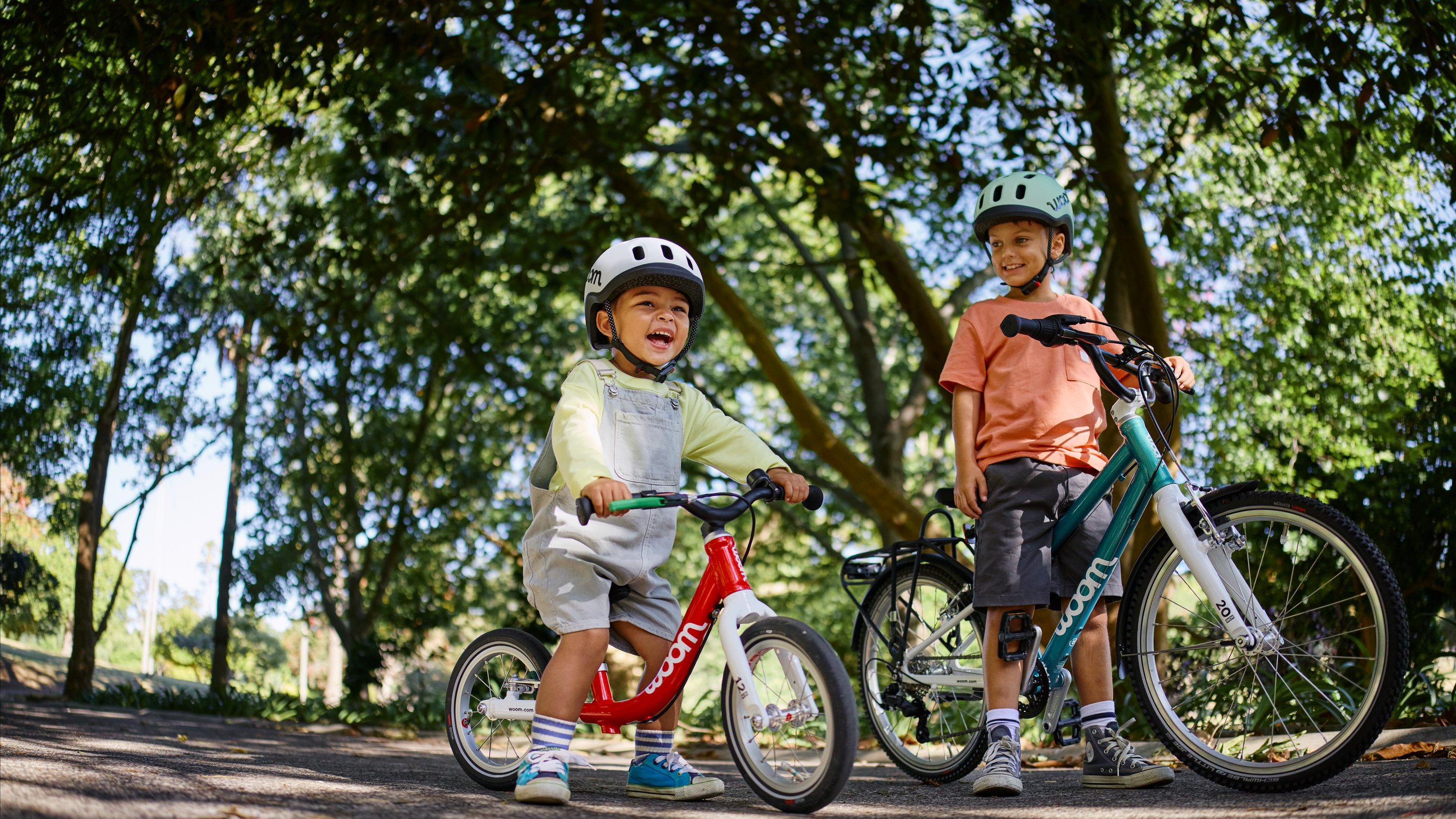 Two children in woom READY kids helmets; one on a red woom GO 1 and one standing by a teal woom GO with a POP basket.